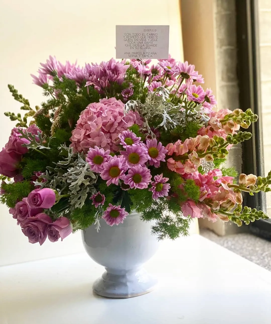 Arreglo de flores rosas y lilas en urna blanca junto a una ventana con luz natural