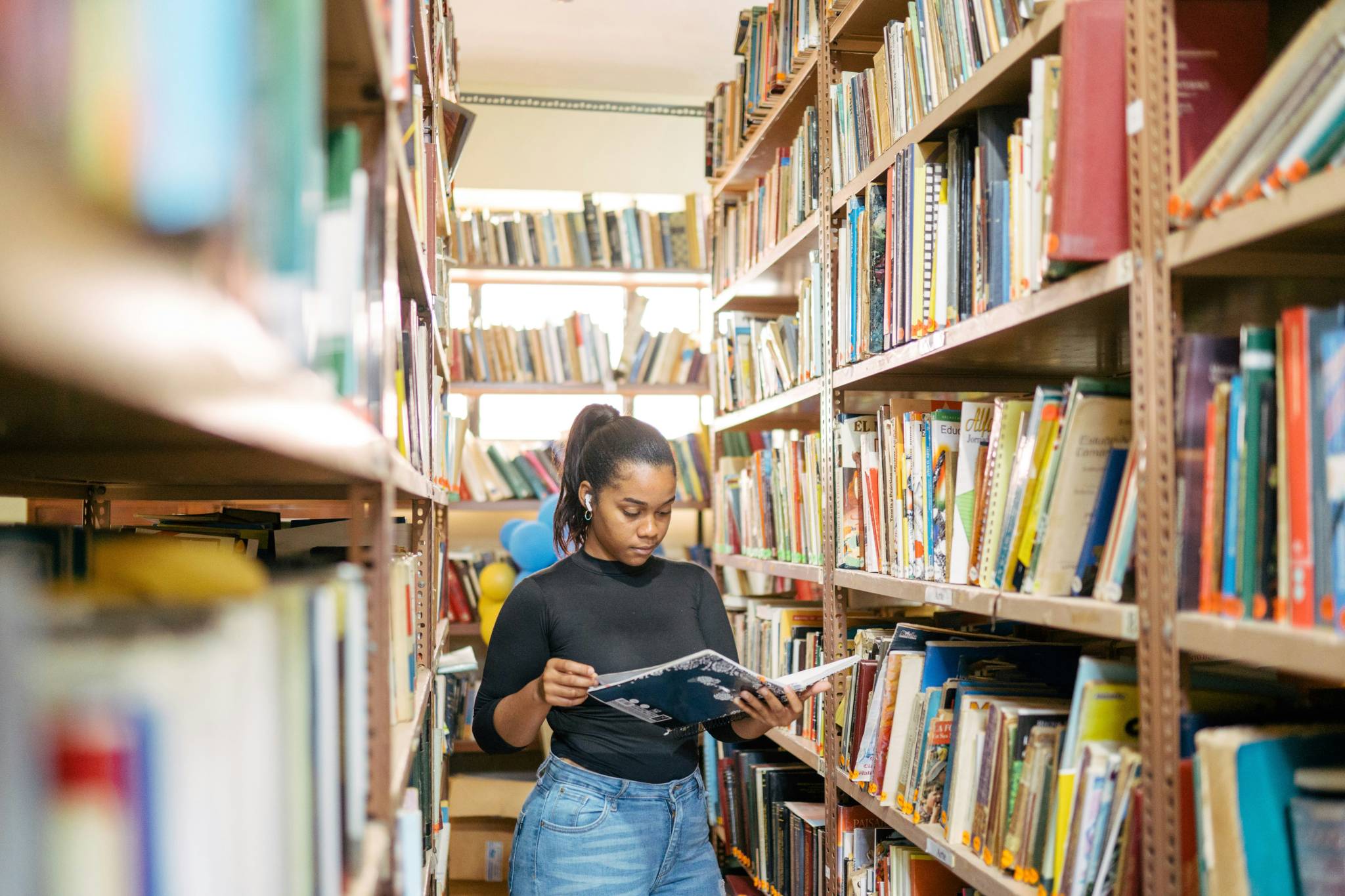 A young woman stands between two tall bookshelves, focused on reading a book in her hands while wearing earbuds. The library is packed with books, and sunlight filters through windows in the background. This scene represents a typical learning environment within the Australian education system.