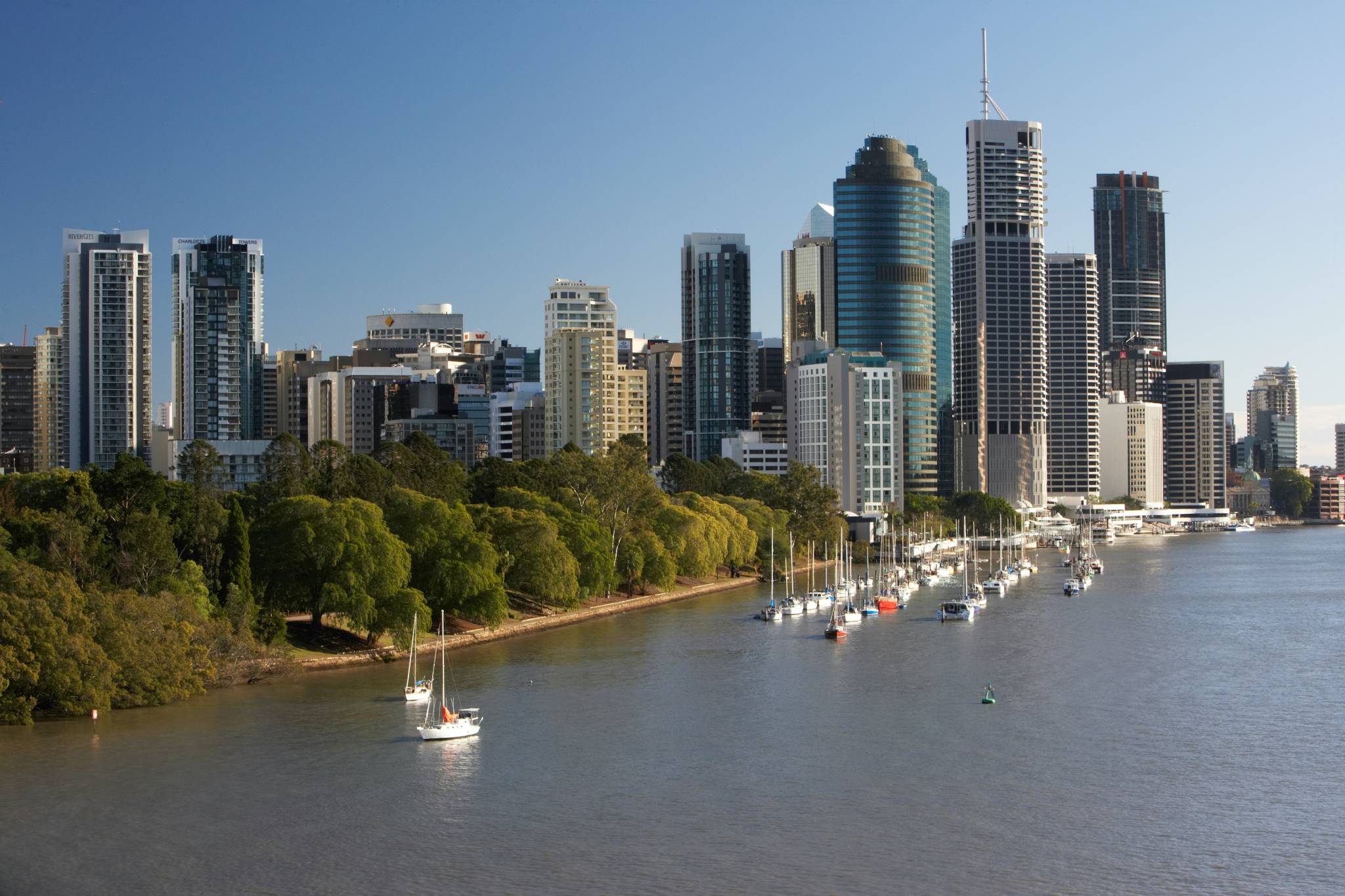 Panoramic view of Brisbane city skyline along the river with yachts anchored by the shore, showcasing the city's modern architecture and riverside lifestyle.