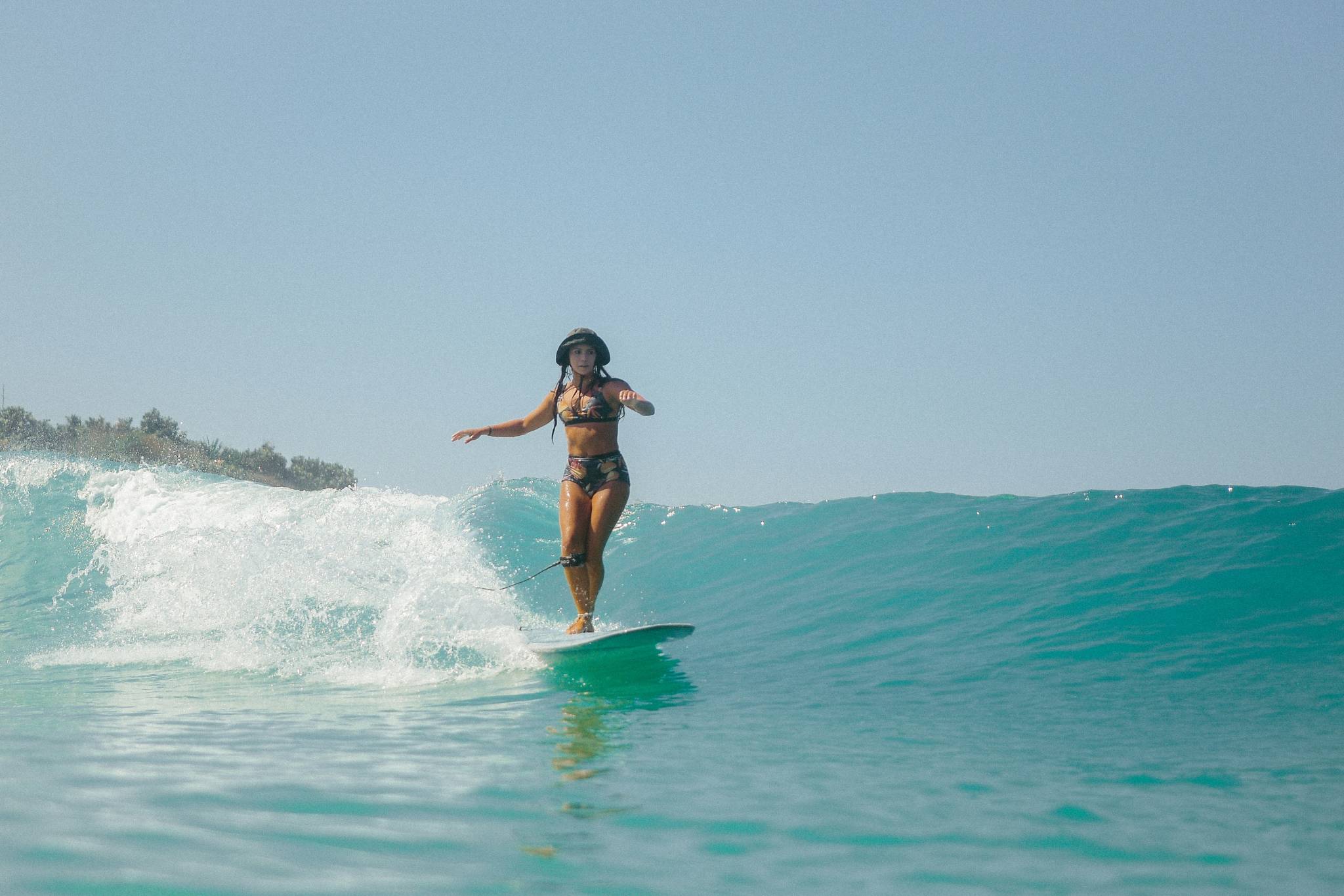 A woman surfing on a calm wave in Byron Bay, Australia, wearing a patterned bikini and a bucket hat, balancing on her longboard under a clear blue sky.