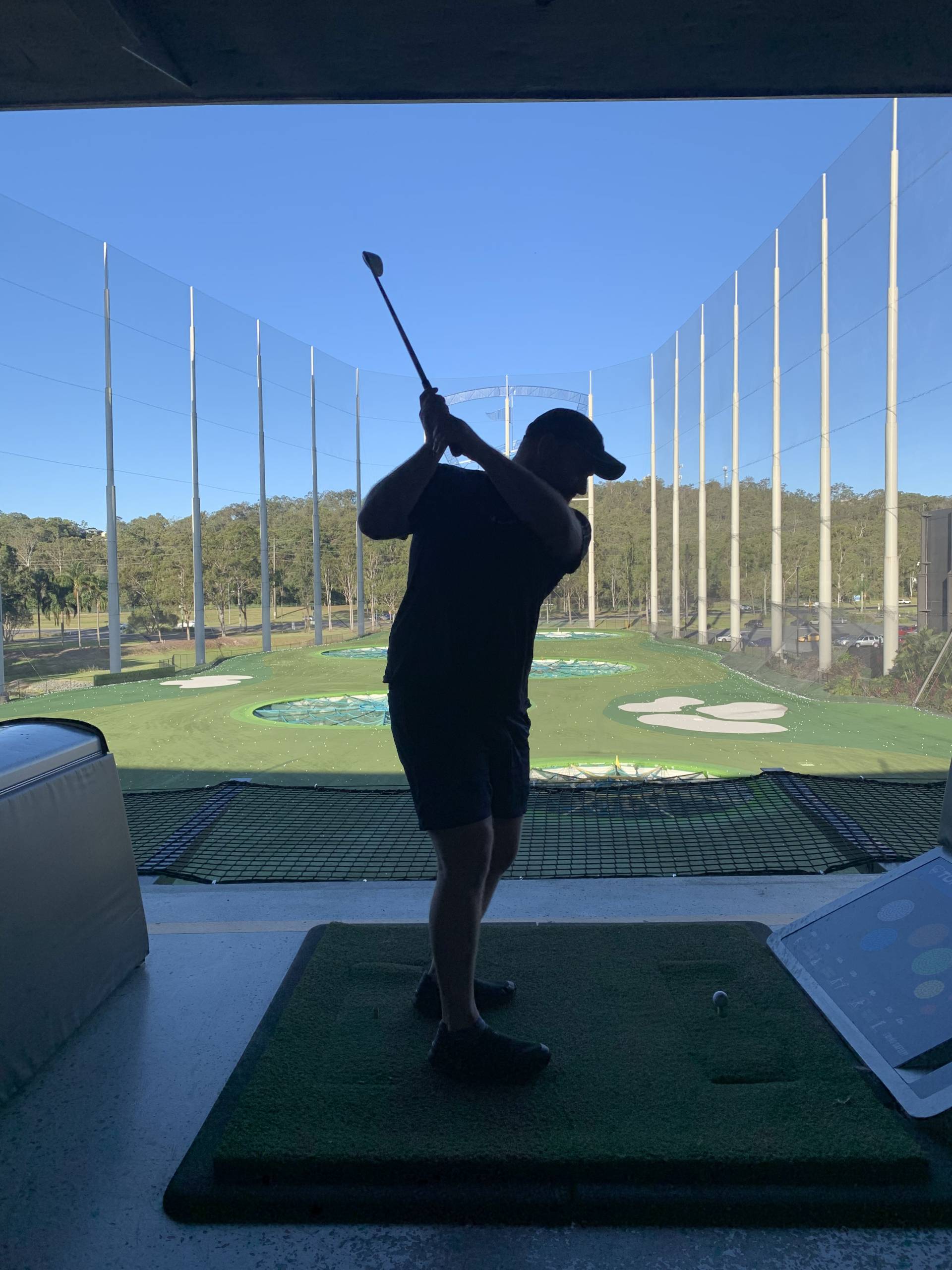 A golfer mid-swing at an outdoor driving range in Brisbane, set against a clear blue sky, a fun activity to try during your Brisbane working holiday.