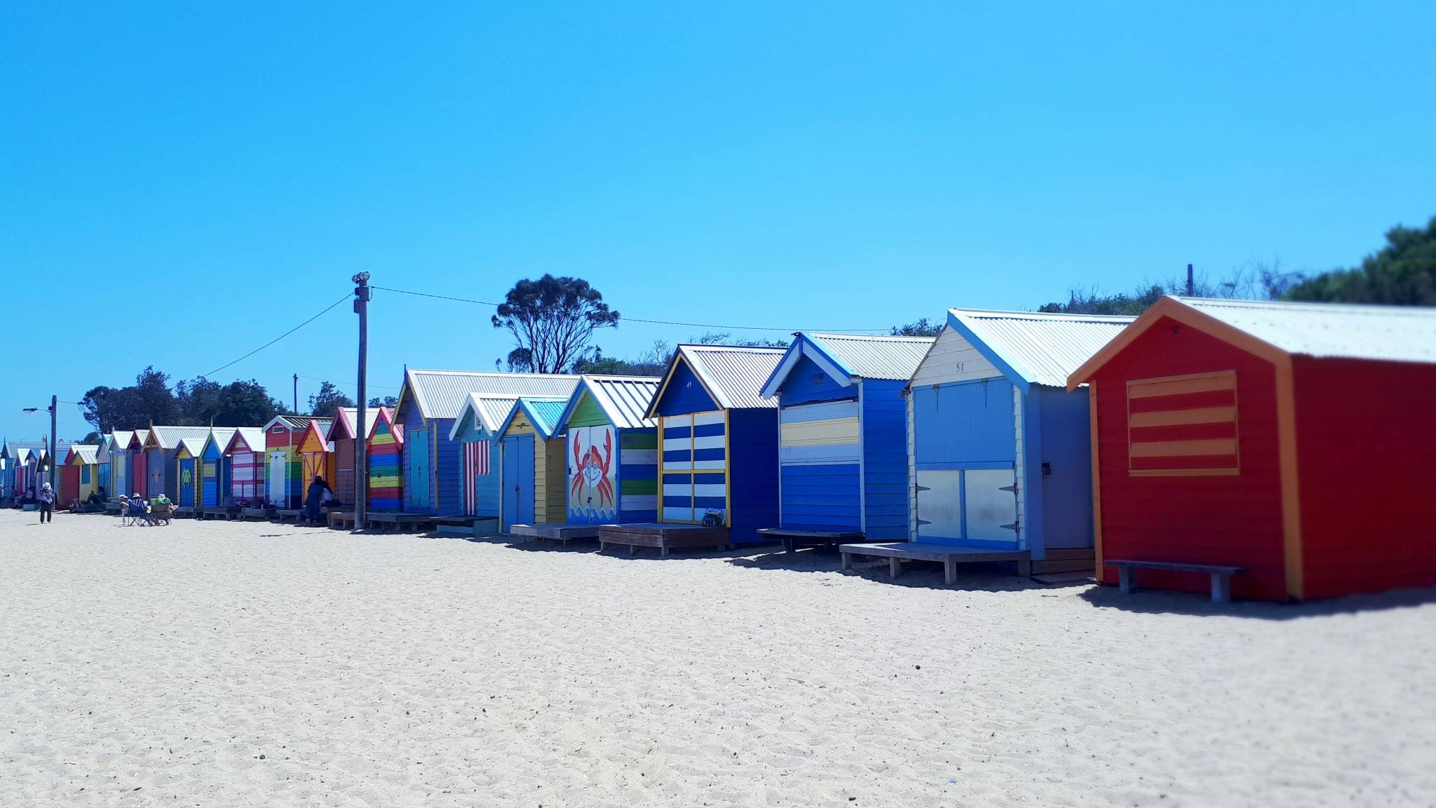 A row of colorful beach huts lined up along a sandy shore under a clear blue sky. Each hut is painted in different vibrant patterns, including stripes, a crab design, and various bright colors.