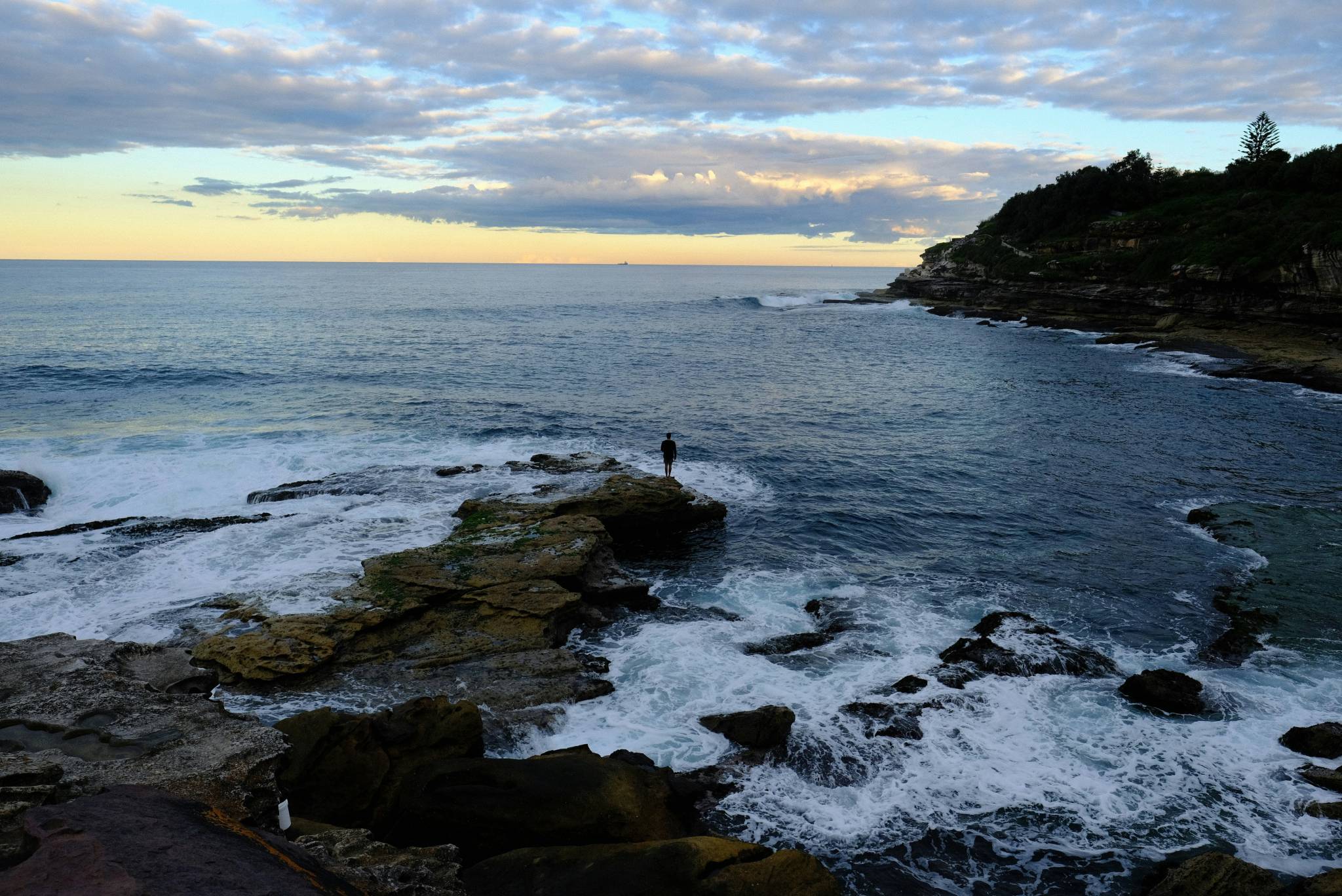 A lone person stands on a rocky outcrop by the ocean, watching the waves crash against the shore at sunset. The sky is filled with soft clouds and the horizon shows a faint glow of sunlight, with cliffs in the distance on the right side of the frame.