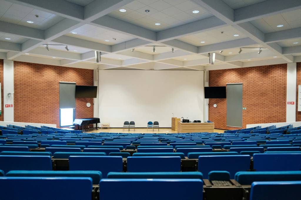 A large, empty lecture hall with rows of blue chairs facing a stage with a white backdrop, podium, and three chairs. The room is well-lit with modern ceiling lights and brick walls.
