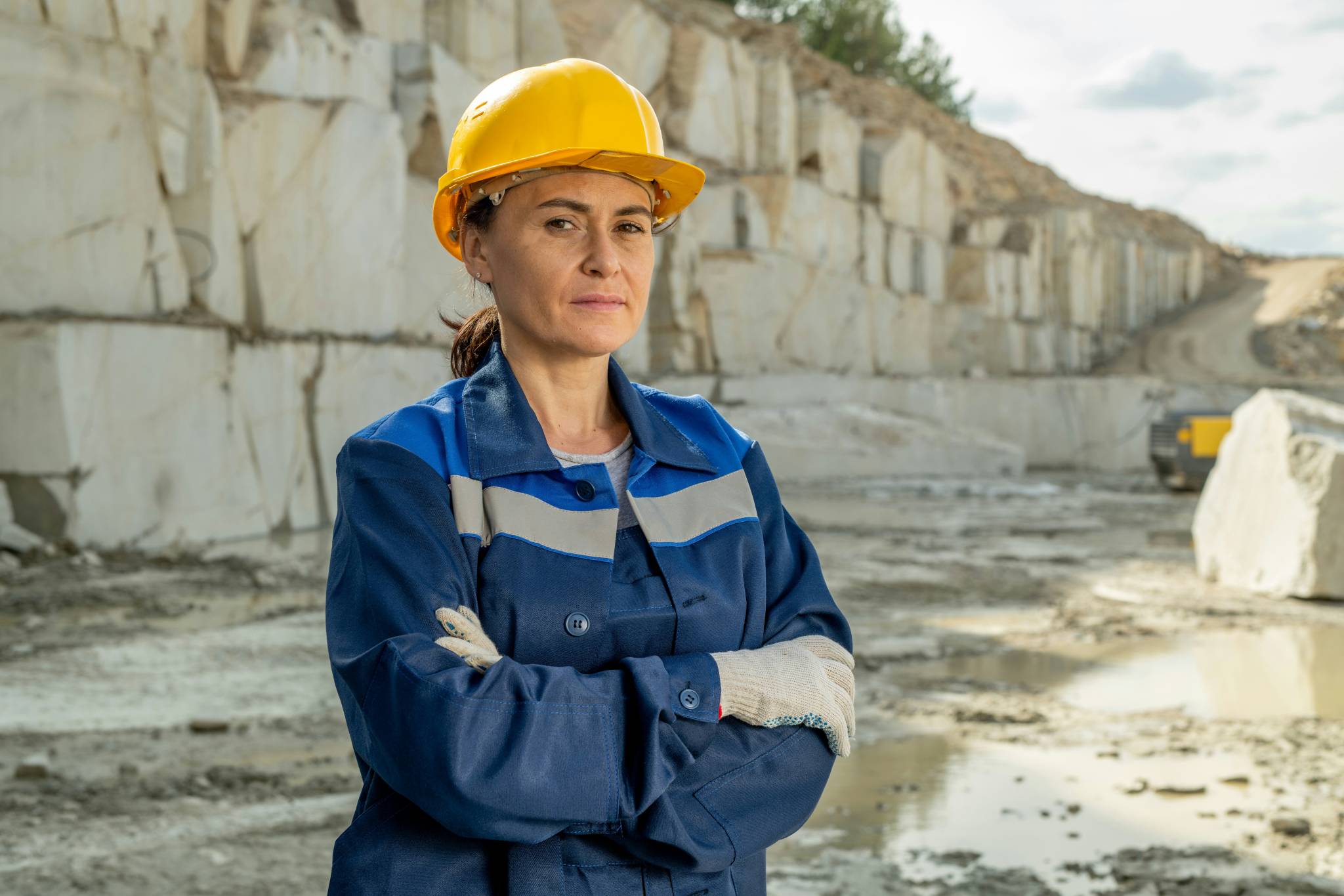 A woman in mining in australia in a yellow hard hat and blue work uniform standing confidently with arms crossed at a quarry site, with rocky, excavated walls in the background.