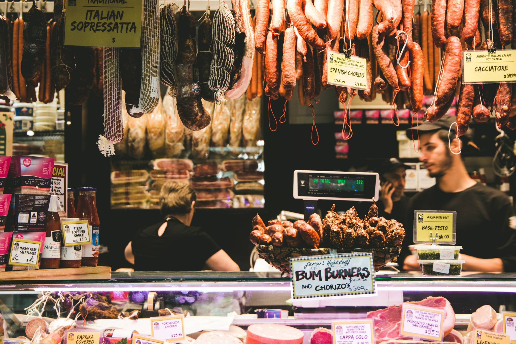 A bustling deli counter at Queen Victoria Market with various types of cured meats, such as salami, chorizo, and soppressata, hanging from the ceiling. Labels like "Bum Burners," "Spanish Chorizo," and "Italian Sopressata" are visible, with staff members working behind the counter.