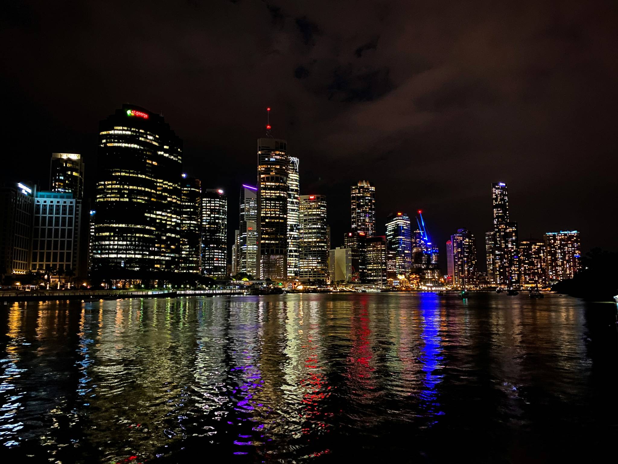 A vibrant nighttime view of Brisbane's city skyline, with illuminated skyscrapers reflecting off the dark waters of the Brisbane River. The buildings glow in various colors, casting vibrant red, blue, and yellow lights across the water. A serene scene for families enjoying free things to do in Brisbane, such as an evening stroll by the river.
