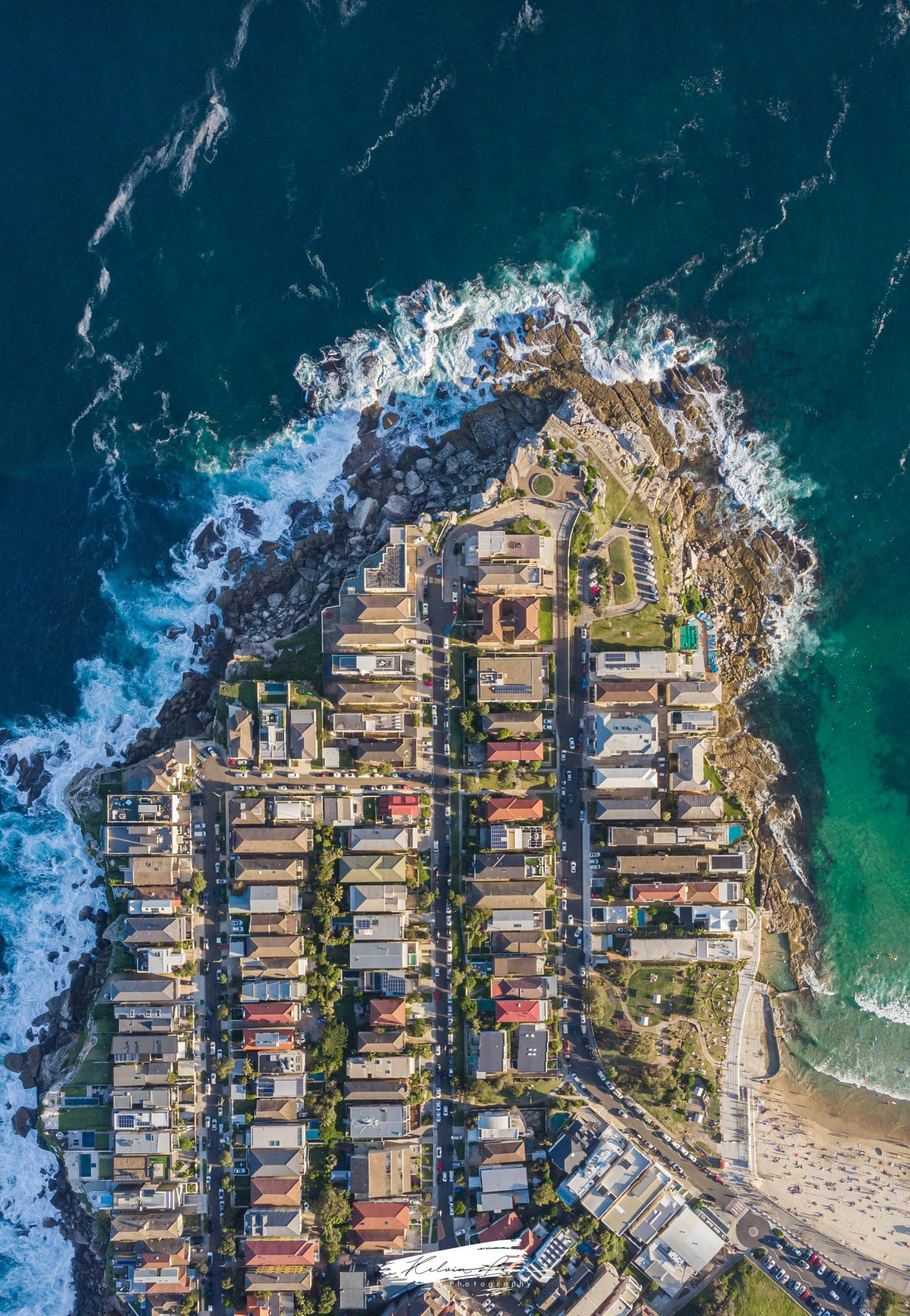 A view of Bondi Beach with gentle waves rolling onto the sandy shore, a distant residential area filled with buildings, and people scattered along the beach. The sky is partly cloudy with shades of blue and white.