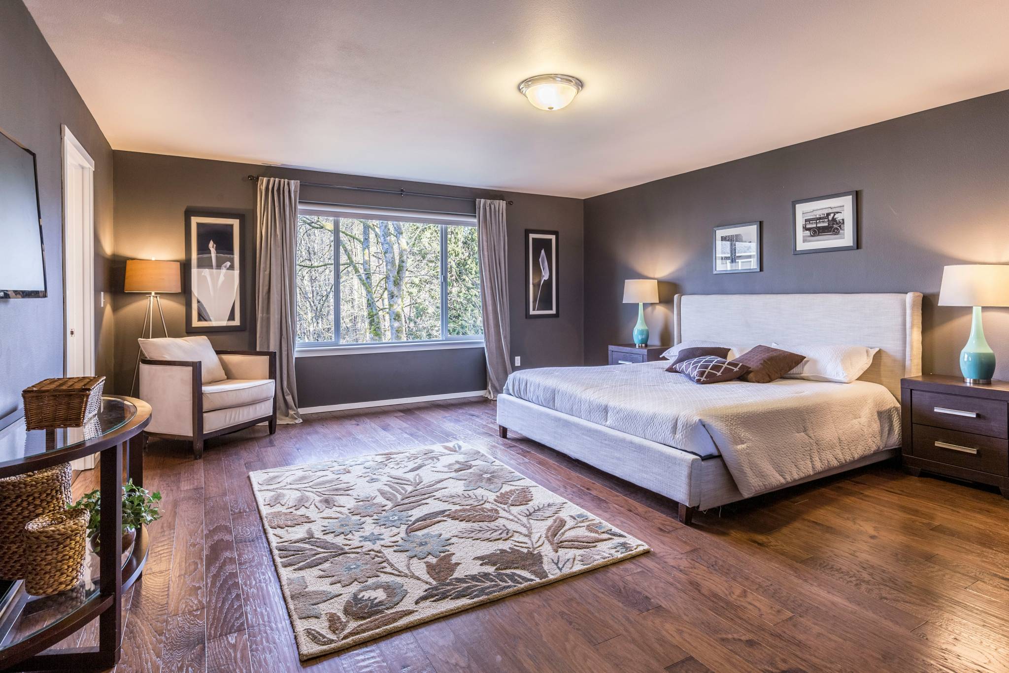 A spacious, modern bedroom featuring a large bed with light gray bedding, flanked by two dark wooden nightstands with teal lamps. The room has hardwood floors, a cream armchair by the window, and a patterned rug with floral designs. A large window offers a view of trees, bringing natural light into the space, complemented by gray walls and minimal decor, including framed art.