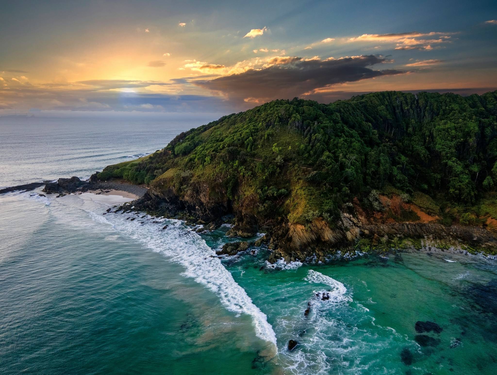 Aerial view of a coastal area in Byron Bay National Park, showcasing a lush green hill, a secluded sandy beach, and clear turquoise waters at sunset.