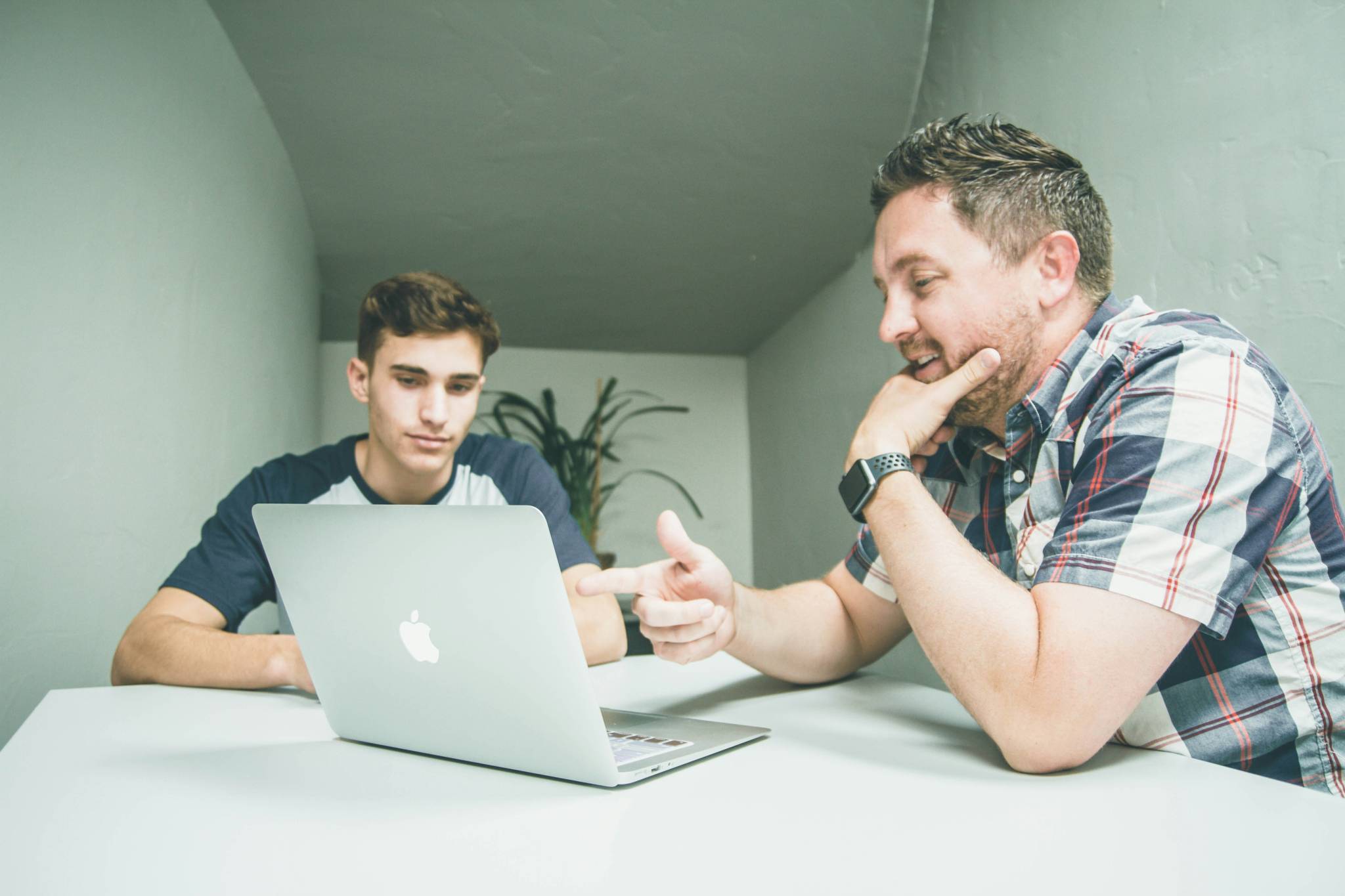 Two men sitting at a table, with one pointing at the screen of a silver Apple MacBook, while the other looks on intently. The scene appears to involve guidance or instruction, with a casual and collaborative atmosphere.