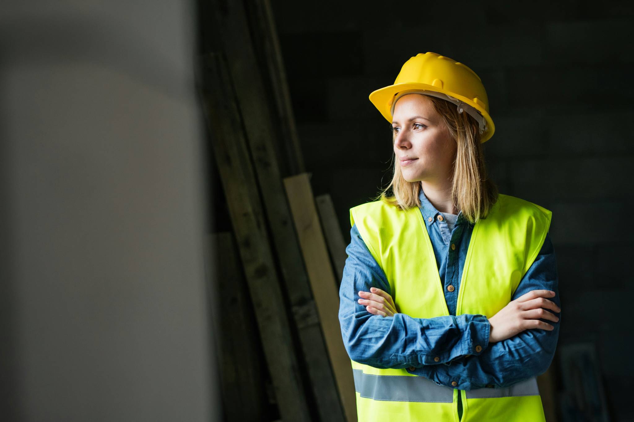 A woman in a construction or mining environment wearing a yellow hard hat and high-visibility vest, standing with arms crossed, looking thoughtfully to the side against a dark background. Keyword: women in mining Australia.