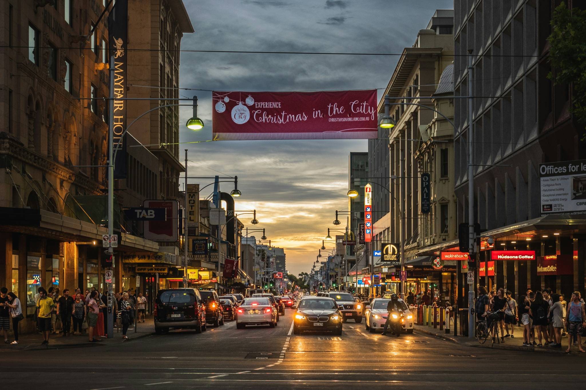 The image shows a busy street in Adelaide at dusk, with cars and pedestrians under a festive banner that reads, "Experience Christmas in the City." The street is lined with lit storefronts, including a prominent Mayfair Hotel sign on the left. The sky is cloudy with a hint of sunset on the horizon, adding to the holiday atmosphere.