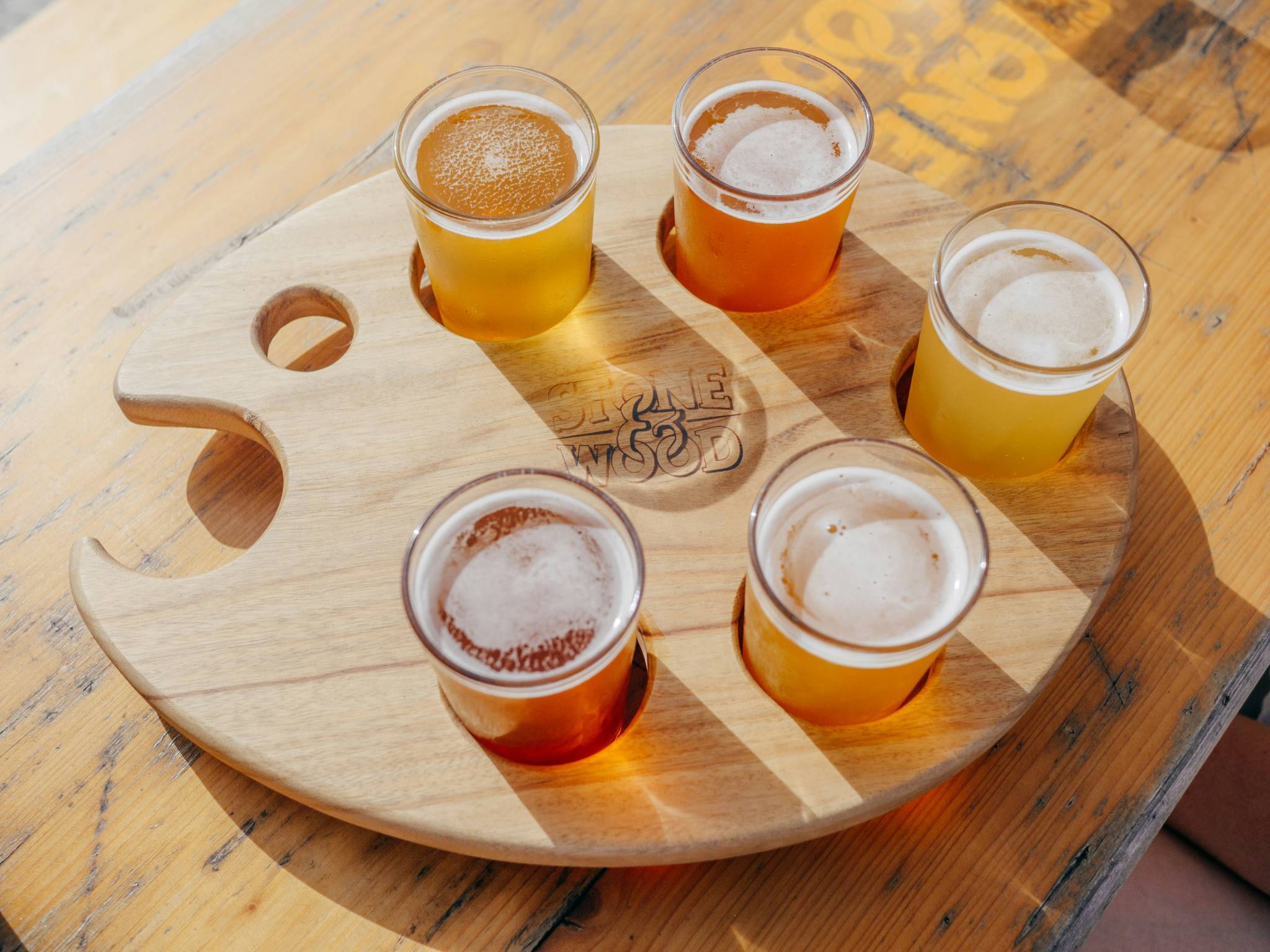 A wooden tasting paddle featuring six small glasses of craft beer from Stone & Wood Brewery in Byron Bay. Each beer varies in color, from light golden to amber, placed on a table with natural sunlight casting soft shadows.