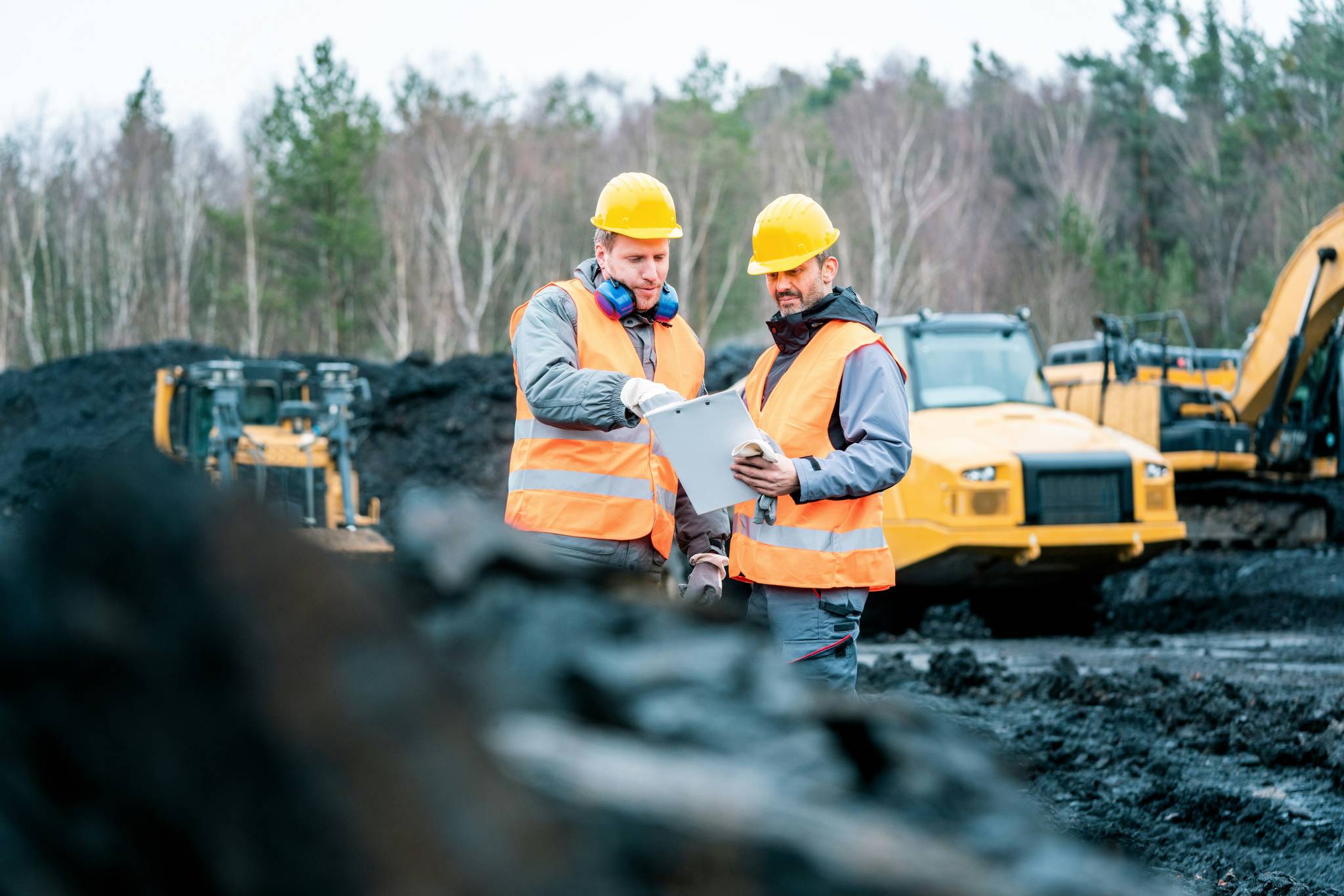 Two men in hard hats and reflective vests reviewing a clipboard at a mining site, with heavy equipment and piles of black material in the background. Making sure that the FIFO mining is not dangerous.