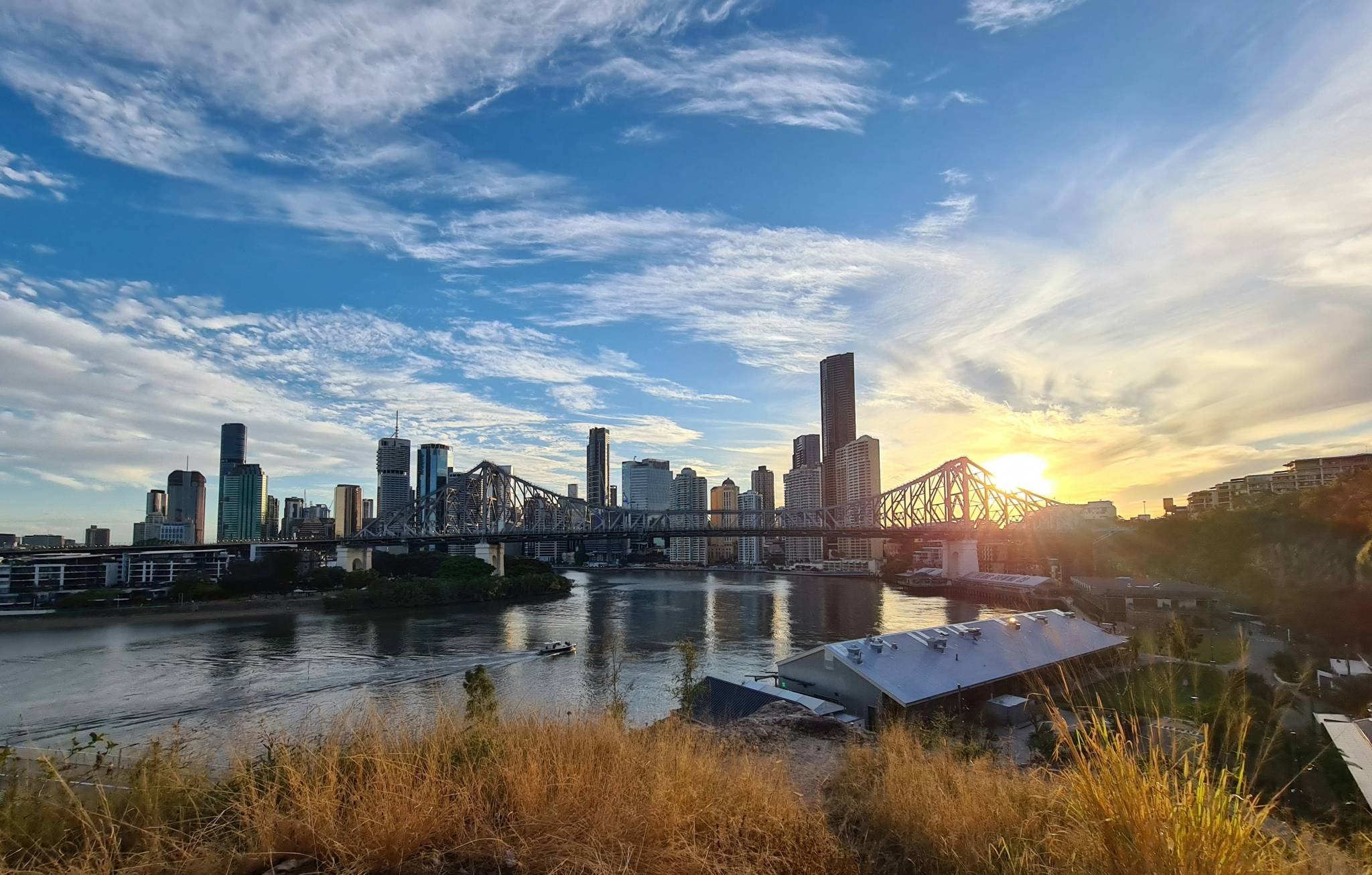 A scenic view of Brisbane at sunset, with the Story Bridge stretching over the Brisbane River, surrounded by modern skyscrapers. A small boat moves along the river, and tall grass is visible in the foreground. This serene scene is perfect for families looking for free things to do in Brisbane with kids, offering a peaceful outdoor experience