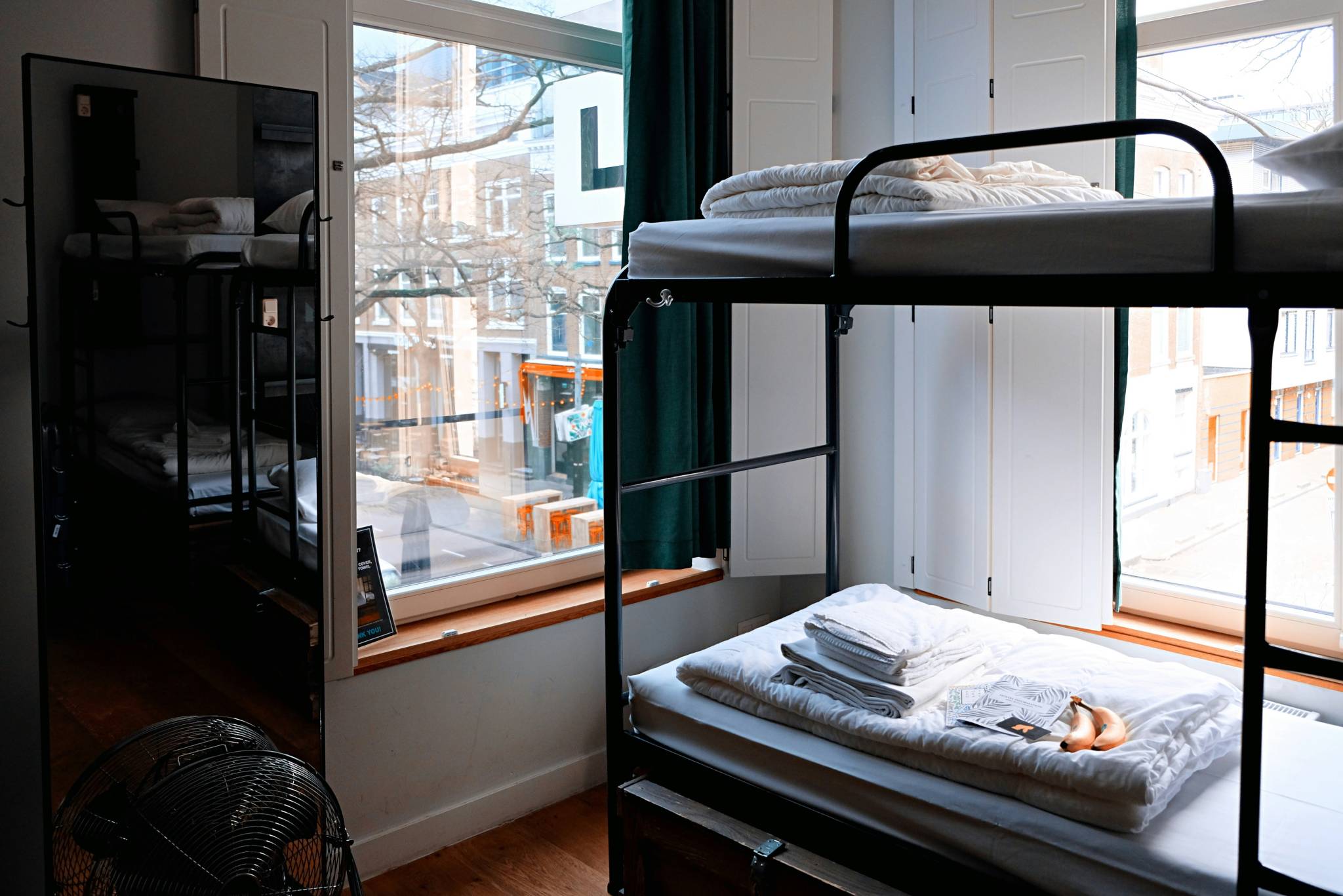 A well-lit room with black metal bunk beds by large windows overlooking a city street. The lower bed is neatly made with folded white linens, towels, a magazine, and bananas on top. A mirror reflects part of the bunk bed, and a small floor fan is visible in the corner.