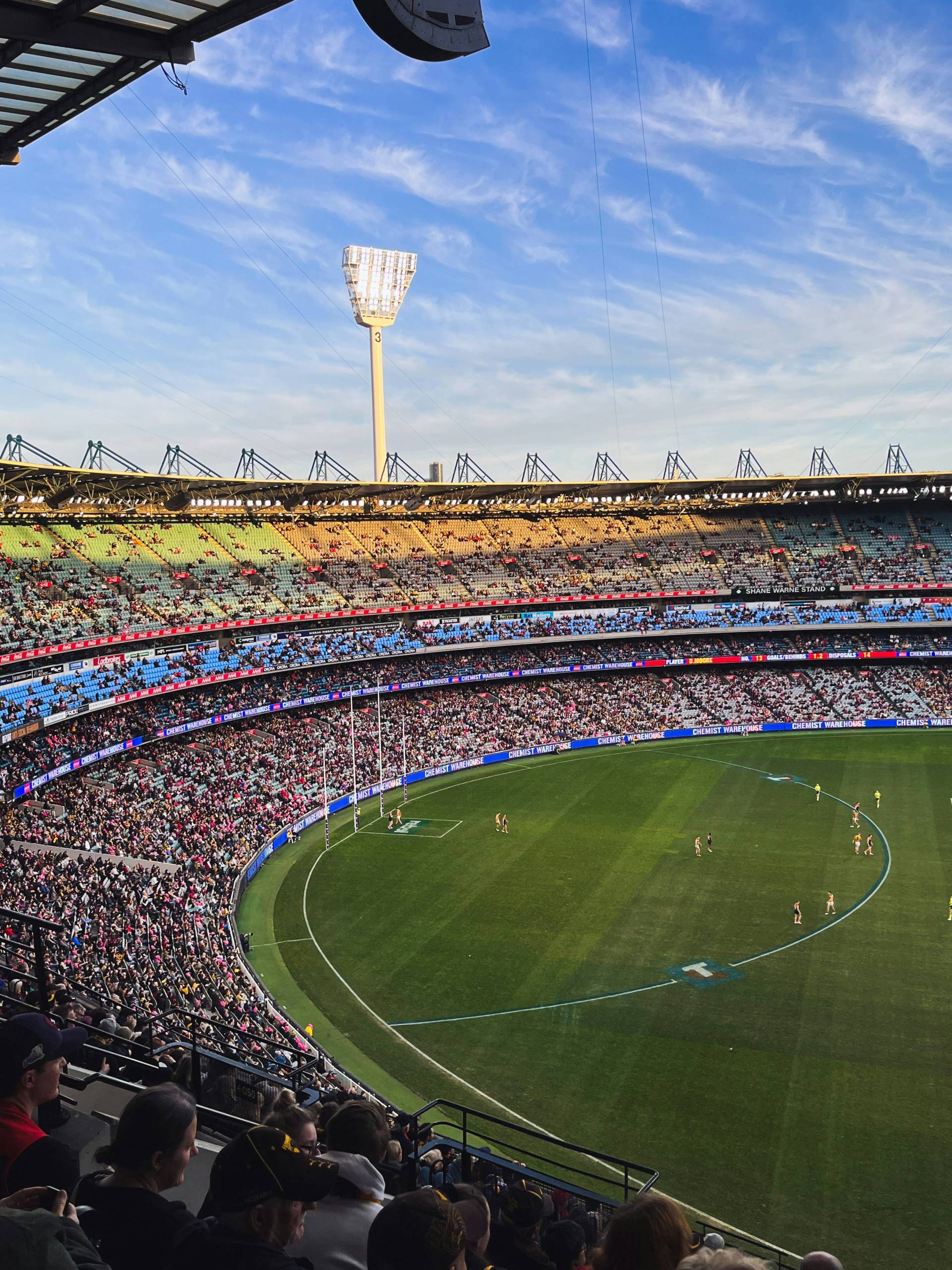 A wide view of a packed crowd at the Melbourne Cricket Ground (MCG during an Australian rules football match. The field is brightly lit under a clear blue sky, with players spread out on the field and spectators filling the stadium.