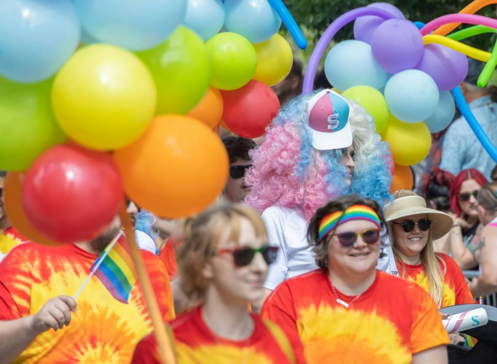 A pride parade filled with participants wearing tie-dye shirts and holding rainbow balloons. A person with a blue and pink wig and a cap stands out among the crowd. Parades like these are part of the diverse and inclusive celebrations that often connect with the community around gay bars in Sydney.