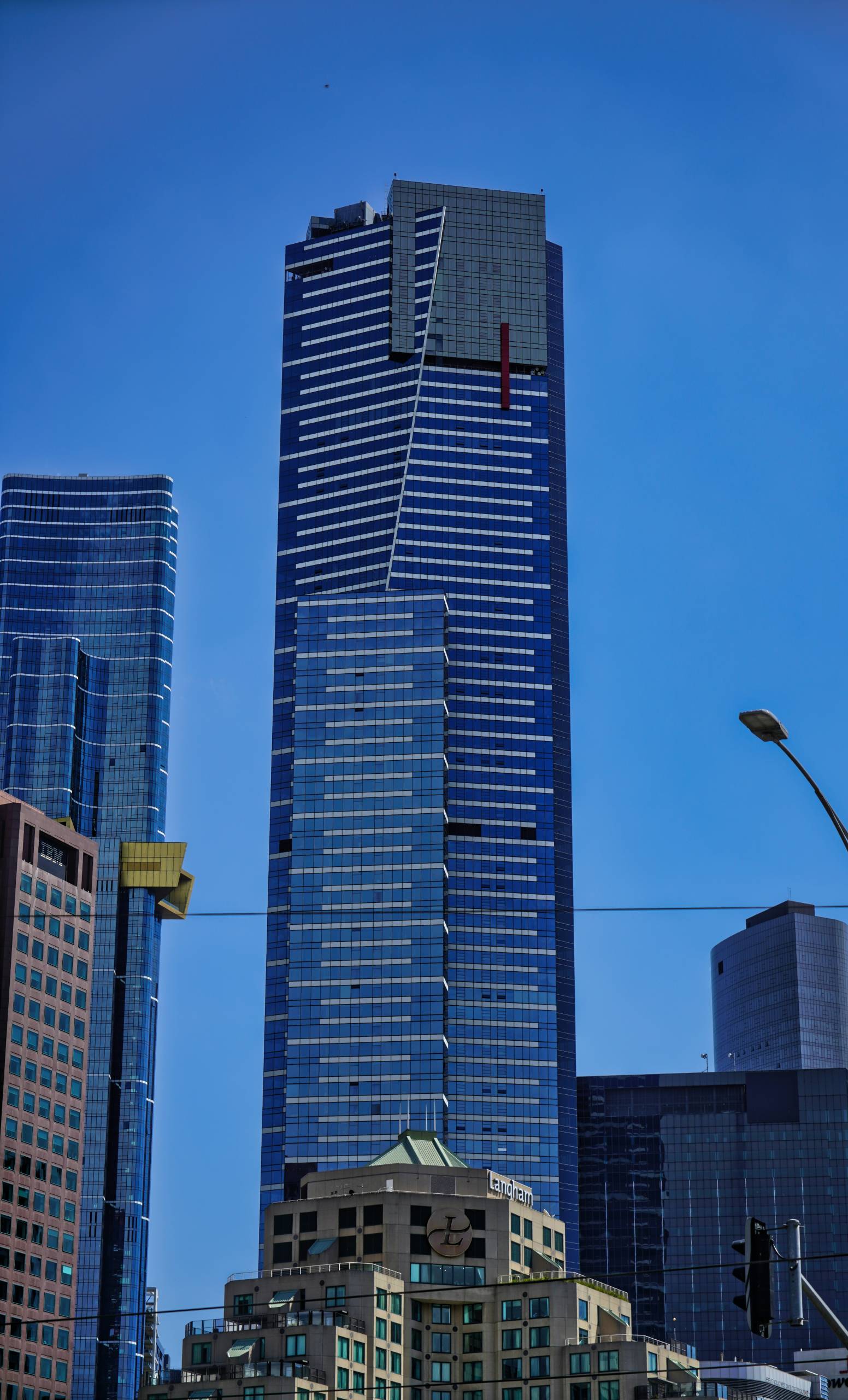A tall modern skyscraper with a sleek blue glass exterior, featuring geometric patterns and sharp lines. Surrounding buildings, including one with "Langham" branding, are visible in the foreground under a clear blue sky.