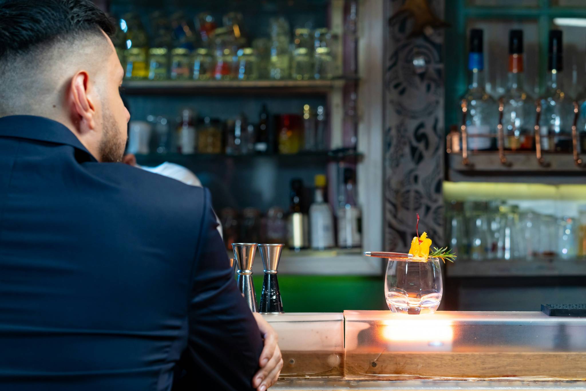 A man in a dark suit sits at a bar, looking at a well-prepared cocktail garnished with fruit and herbs. Gay bars in Sydney provide stylish, upscale experiences for their patrons, complete with carefully crafted cocktails and a modern setting.
