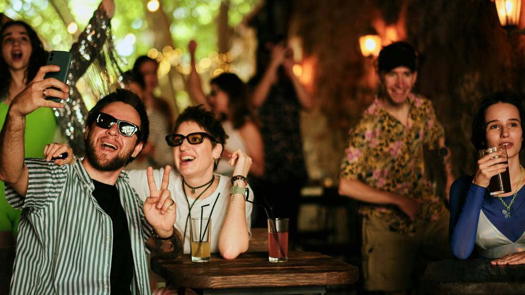 A group of friends, two in the foreground making peace signs, taking a selfie while celebrating at a lively bar. Gay bars in Sydney often offer an energetic and vibrant atmosphere like this, where people gather for drinks and fun.