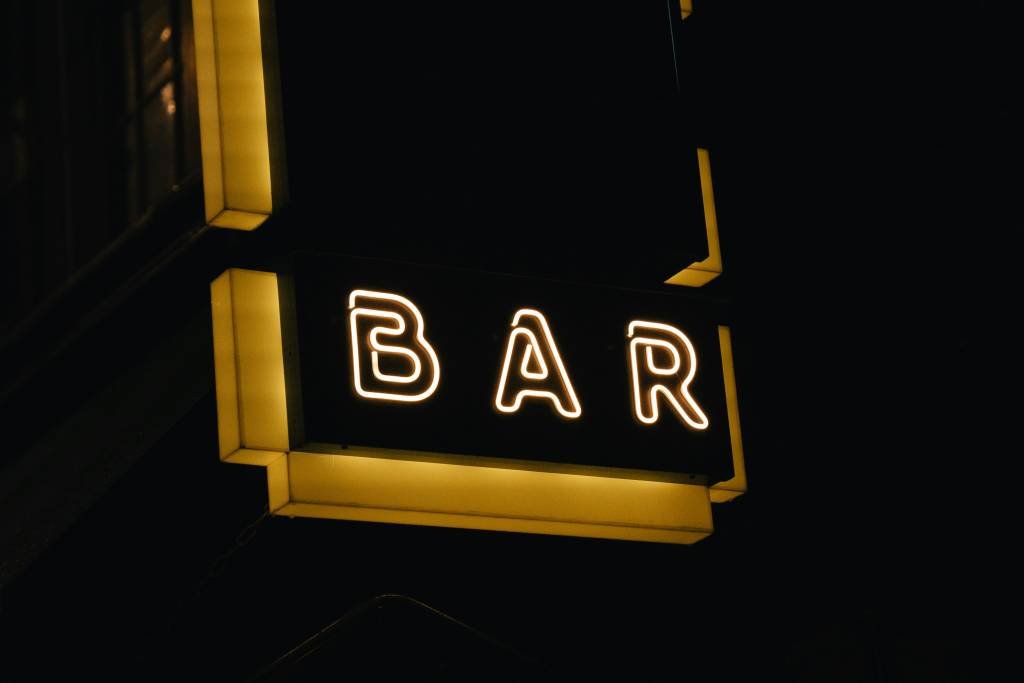 A glowing yellow neon sign with the word "BAR" against a dark background. This simple and sleek sign can be found in many gay bars in Sydney, symbolizing a warm and welcoming atmosphere for nightlife