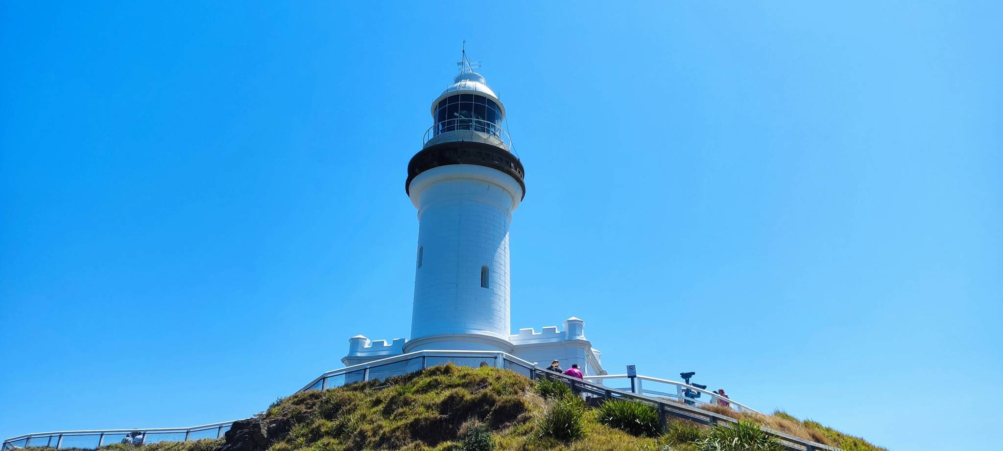 The image shows the Byron Bay Lighthouse in New South Wales, Australia, standing on a grassy hill beneath a bright, clear blue sky. The iconic white structure, known for marking Australia's easternmost point, has a railing near its base where visitors can be seen enjoying the scenic coastal views.