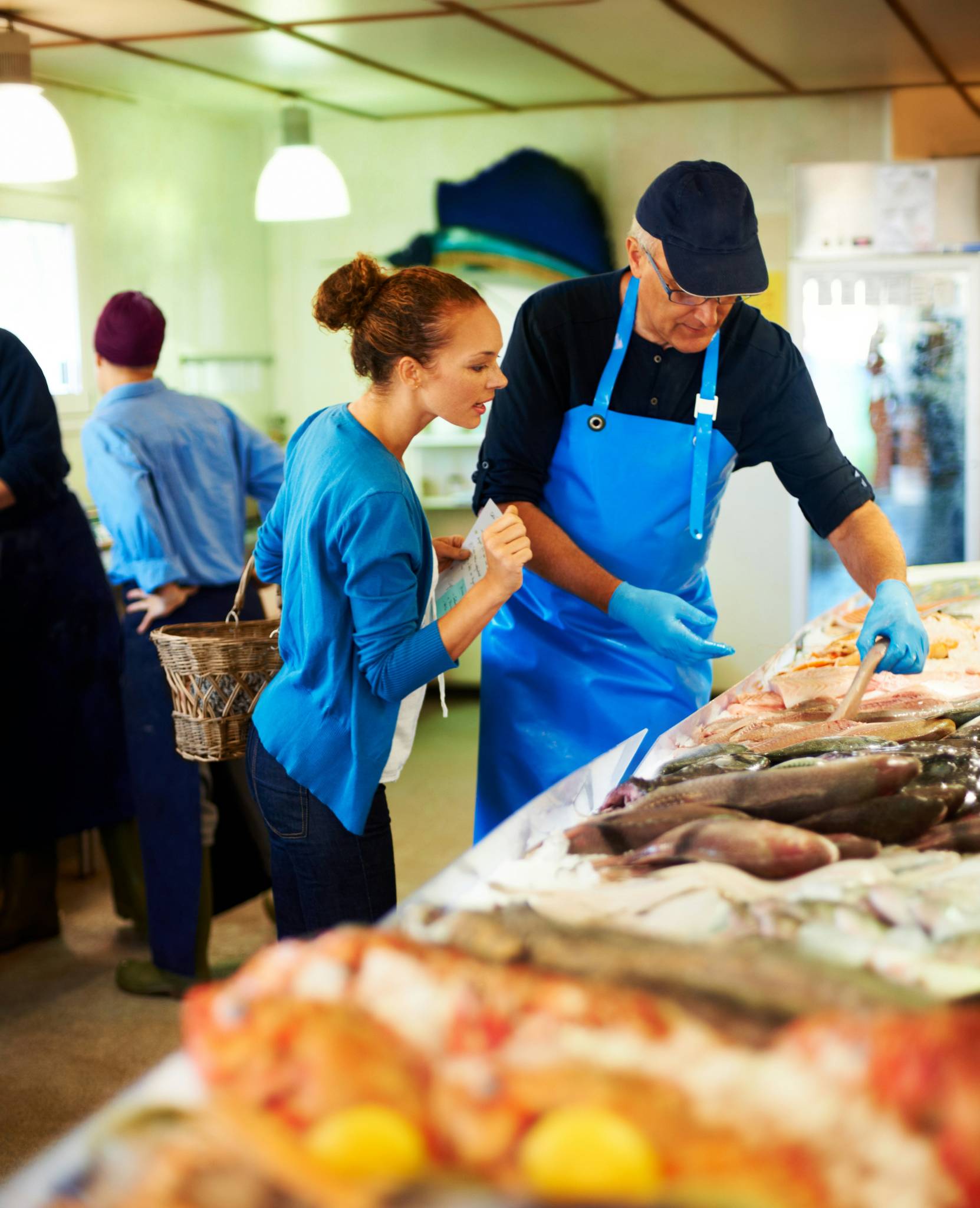 A woman shopping at a fish market in Sydney, guided by a fishmonger wearing a blue apron and gloves, who points out various fresh seafood options. This bustling market experience highlights a fun and interactive activity to do with friends in Sydney, especially for food lovers exploring local cuisine. This could be a Australian backpacker job.