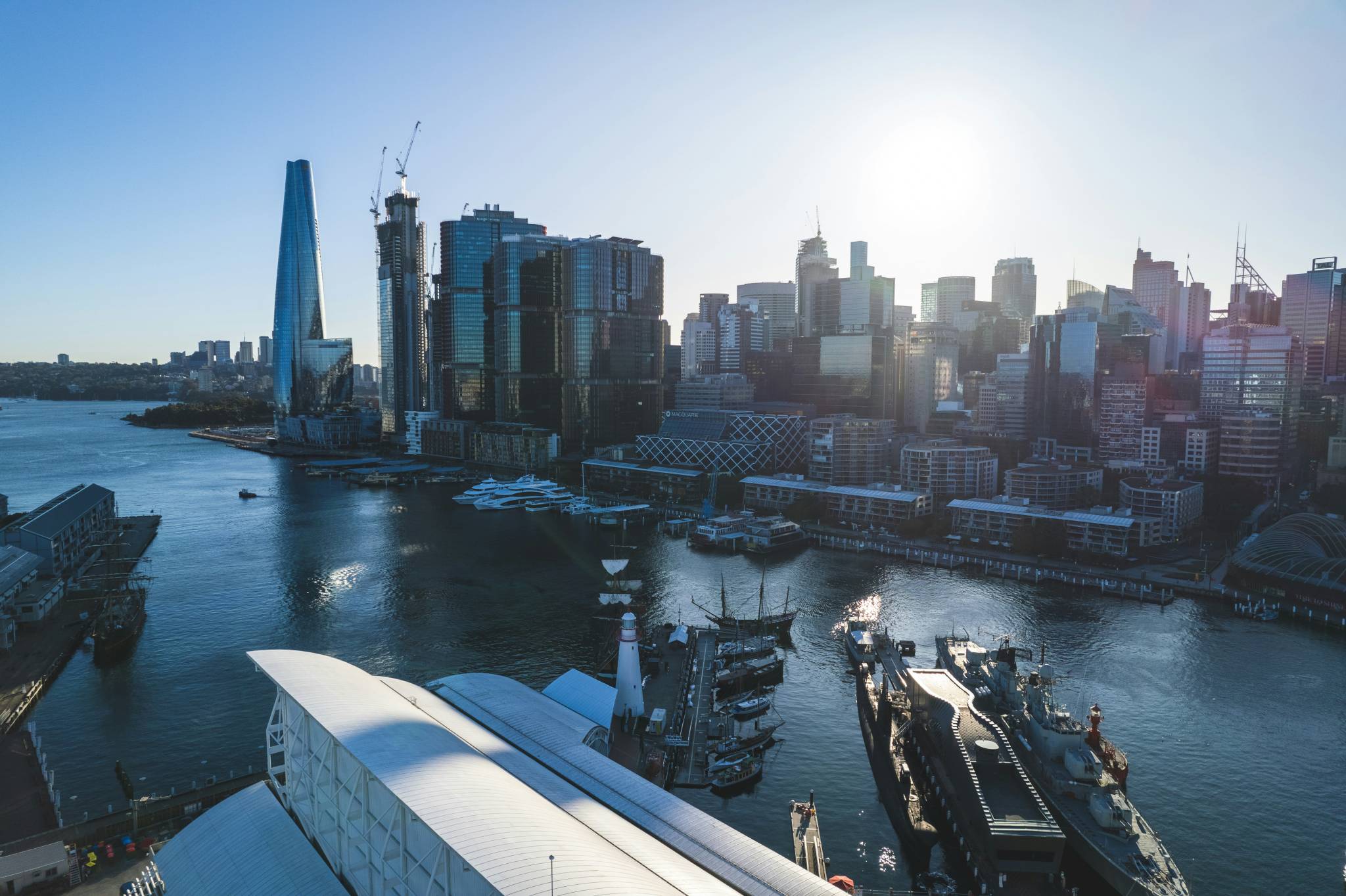 A sunny aerial view of Sydney's harbor featuring modern skyscrapers, including the Crown Sydney building, with yachts and ships docked in the blue waters. The city skyline glows under the bright sun, an ideal spot for exploring the best backpacker bars in Sydney.