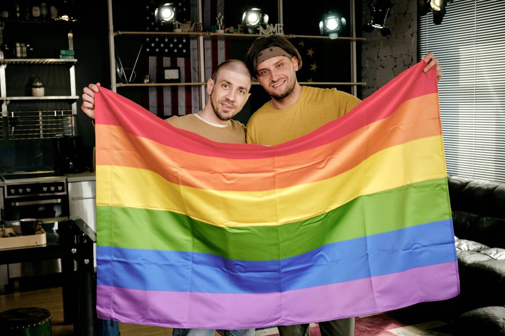 Two smiling men, one bald and the other wearing a bandana, hold up a large rainbow flag, symbolizing LGBTQ+ pride. This celebration of diversity is often a common sight in gay bars in Sydney, where inclusivity is embraced.
