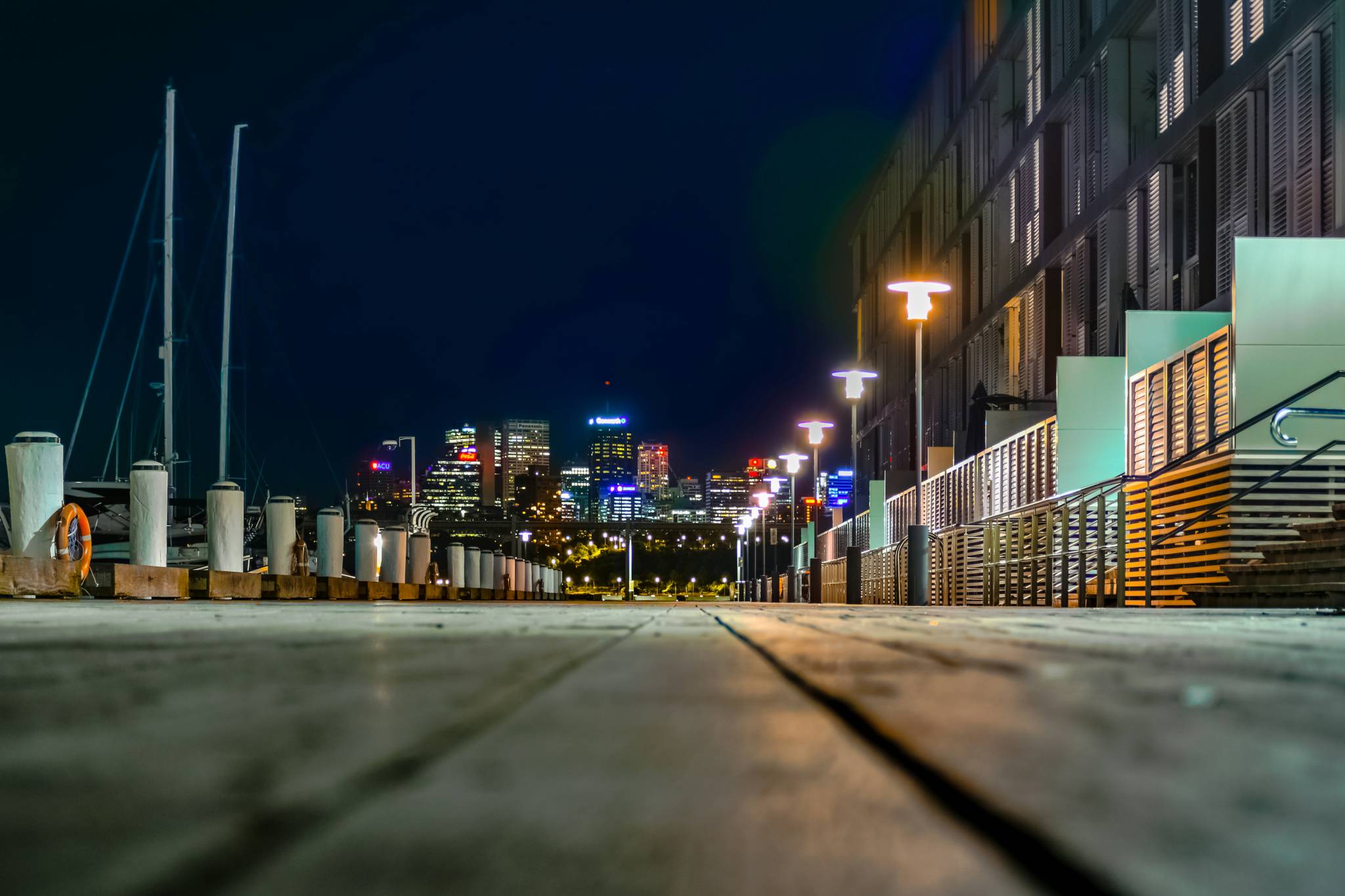 A nighttime shot of a wooden boardwalk leading towards a distant, illuminated city skyline. The warm street lamps guide the way, making this a great area for those looking to experience the best backpacker bars in Sydney.