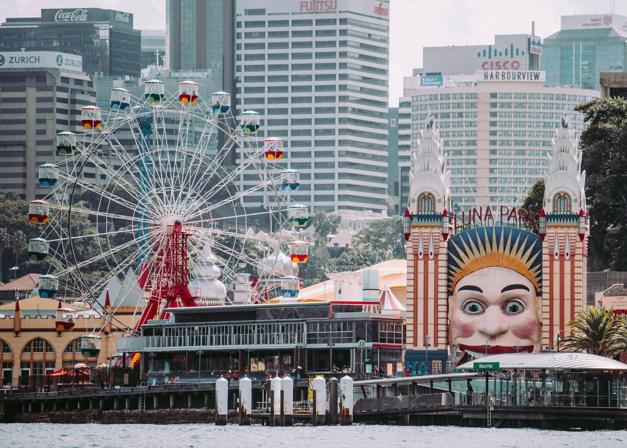 Sydney's Luna Park entrance with its iconic smiling face facade and tall, colorful Ferris wheel in the foreground, set against a backdrop of modern skyscrapers, including logos from companies like Zurich, Coca-Cola, and Cisco. The waterfront location adds a touch of vibrant city-meets-playground charm.