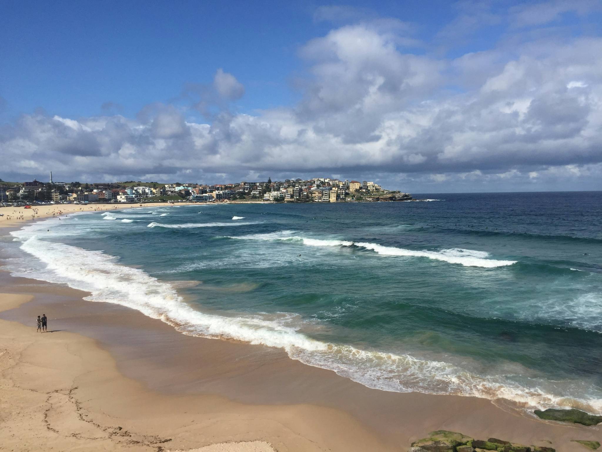 A view of Bondi Beach with gentle waves washing up on the sandy shore. The coastline curves toward a distant residential area filled with buildings, while people are scattered along the beach under a partly cloudy sky.