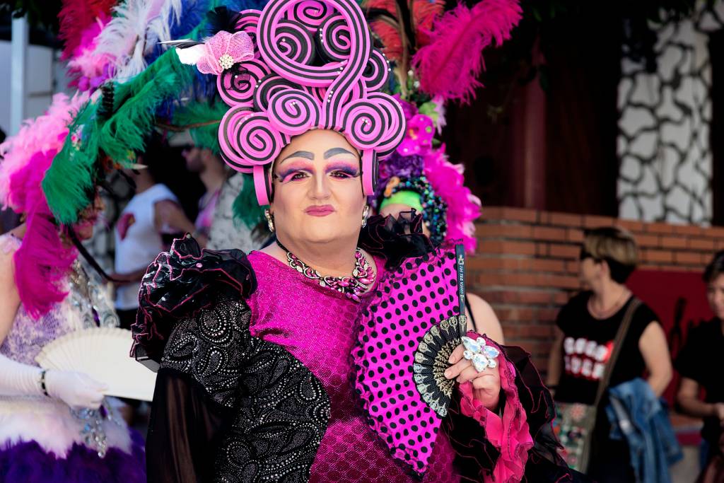 A drag performer wearing a bright pink and black outfit with an intricate headpiece, holding a polka-dotted fan. Events like drag shows and LGBTQ+ parades, commonly associated with the vibrant culture of gay bars in Sydney, often feature such colorful performances.