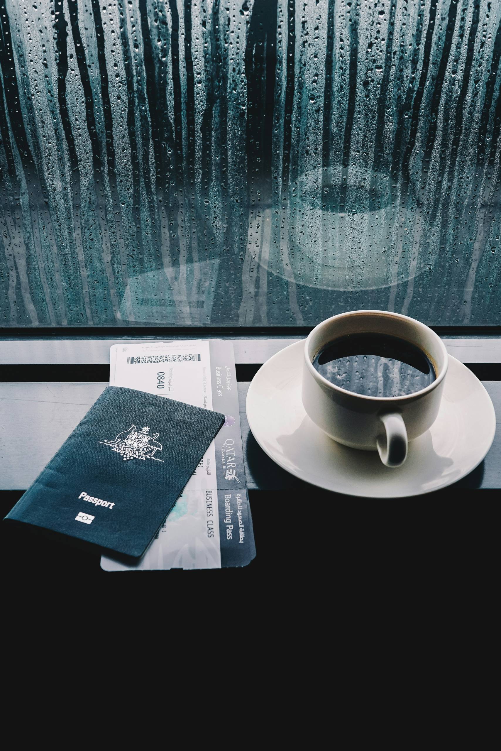 An Australian passport and a boarding pass for Qatar Airways business class lie on a table next to a cup of black coffee. The scene is set against a rain-streaked window, evoking a sense of quiet anticipation before a journey.
