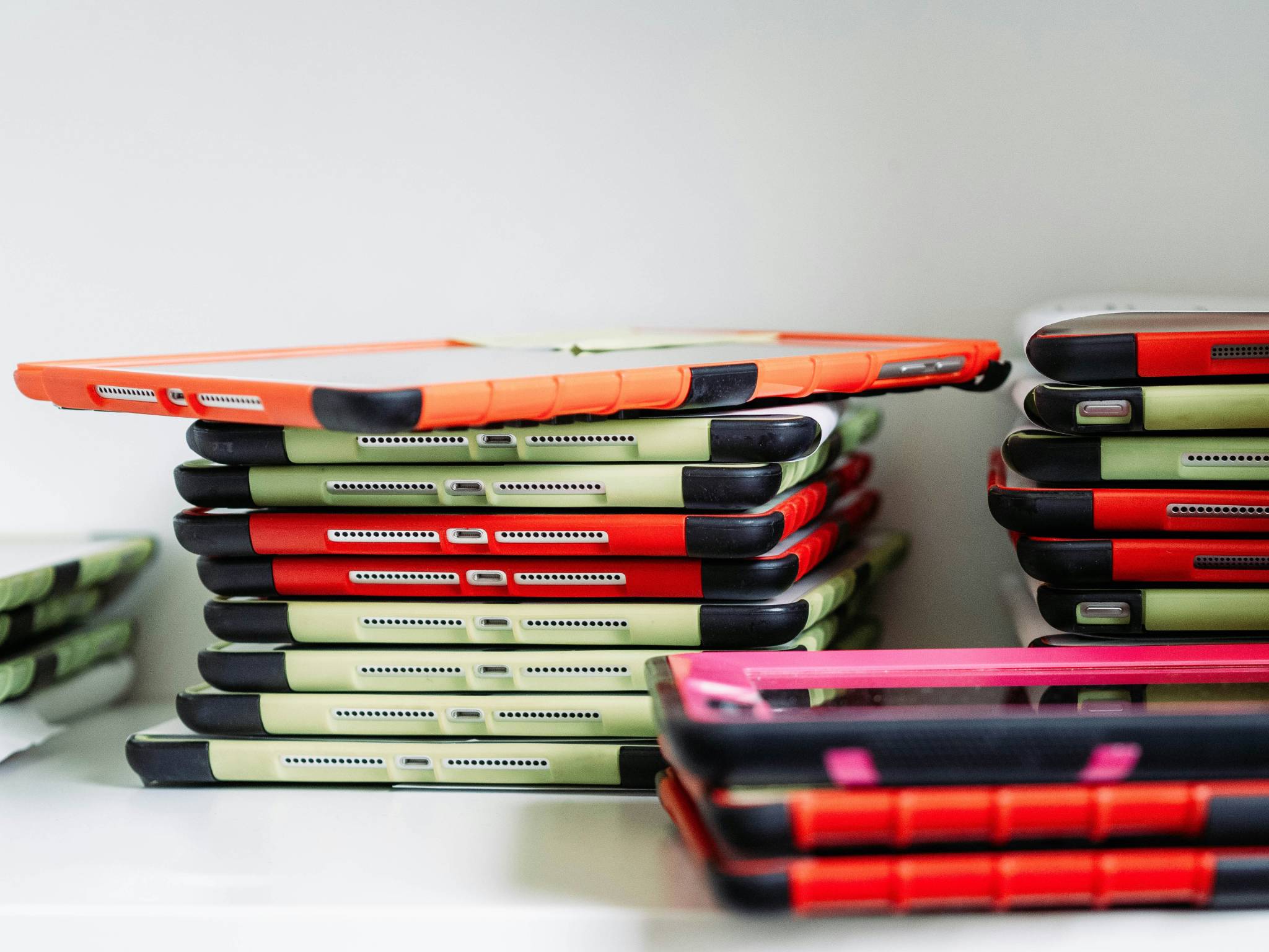 A stack of brightly colored tablets with protective cases in red, green, and black, neatly piled on a shelf. The devices appear ready for classroom use. Teaching in Western Australia.