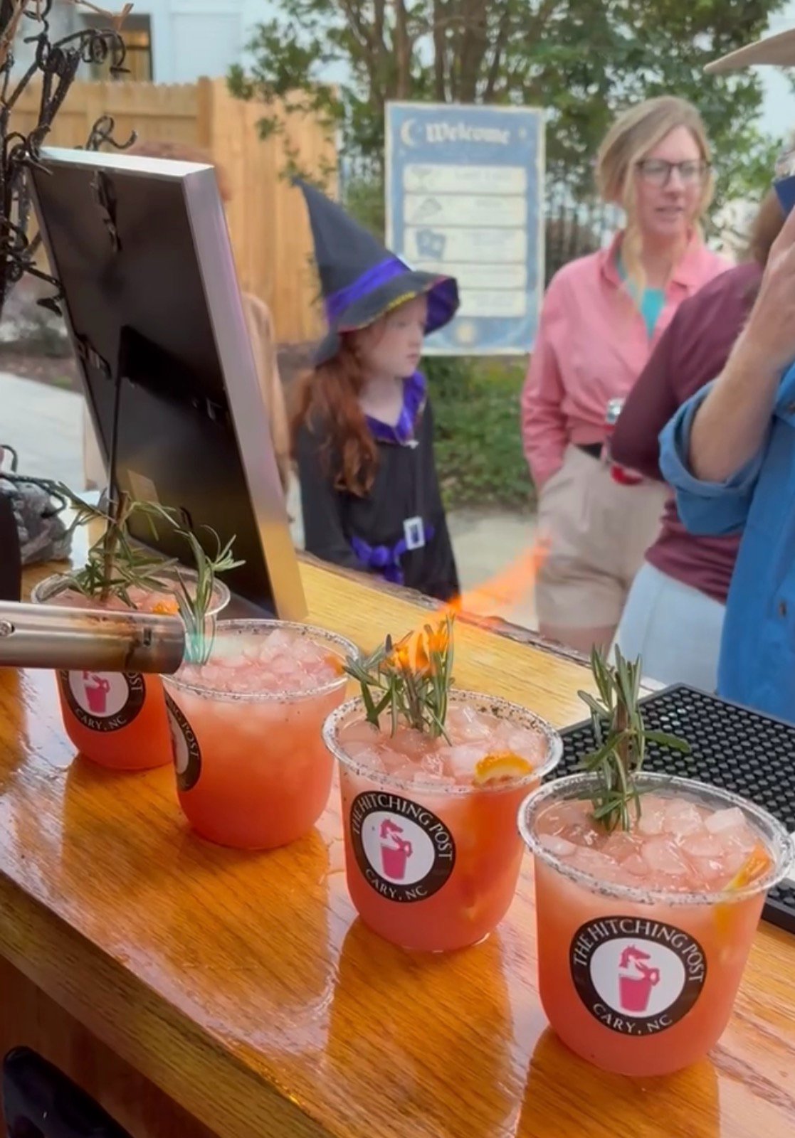 Hitching Post cocktails with torched rosemary garnishes on the bar at a Halloween event, with a young guest in a witch hat in the background