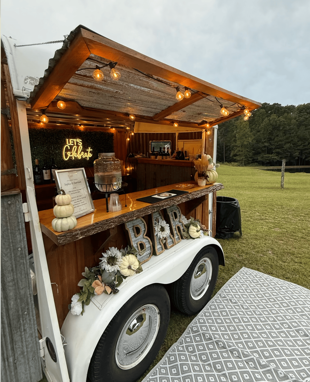The Hitching Post trailer set up on a grassy farm field with a pond, fall pumpkins, sunflowers, and a custom Sophia and Harrison wedding menu