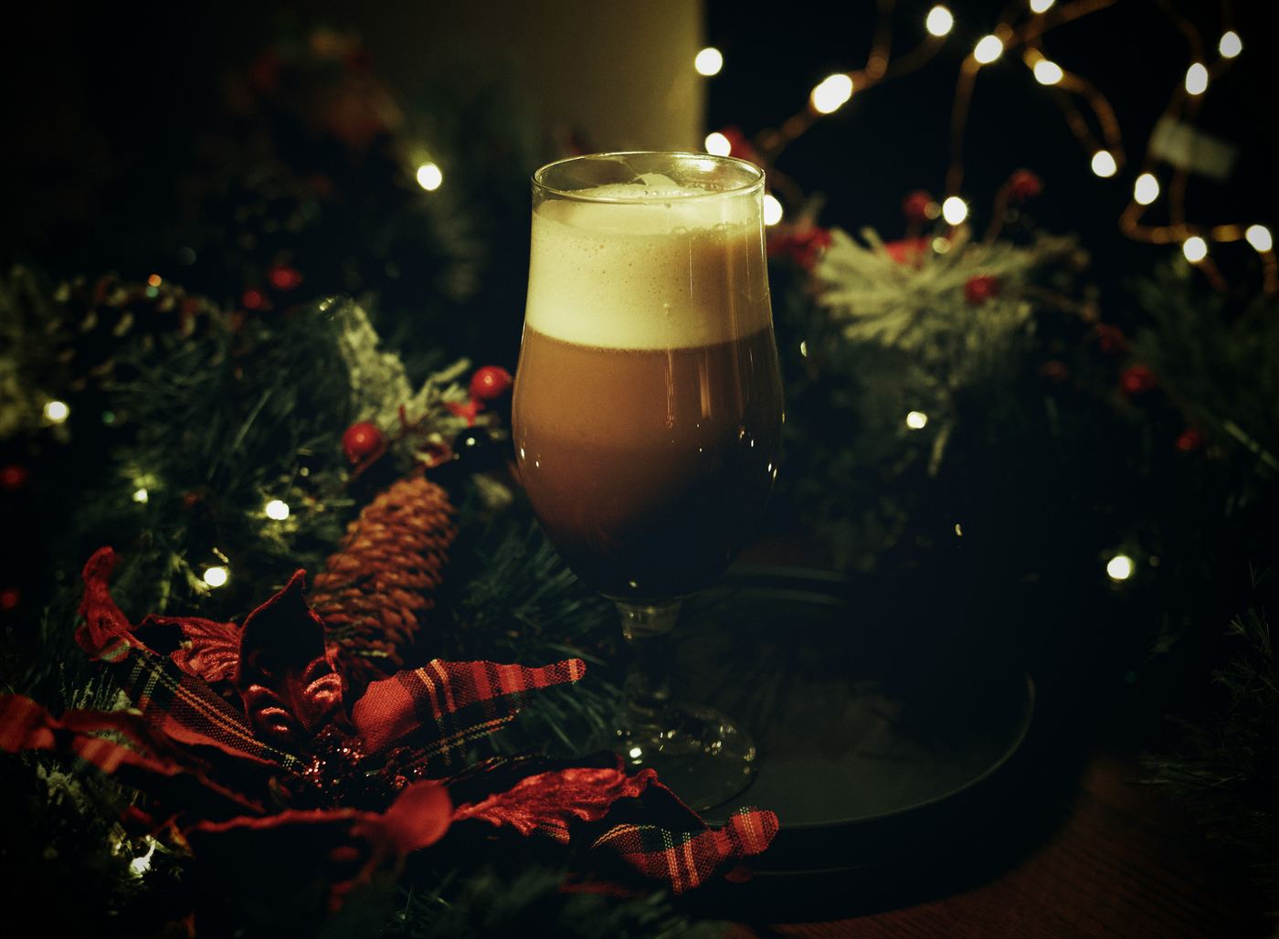 Glass of dark Norwegian Christmas beer with a foam head, surrounded by Christmas lights, pine branches and red decorations