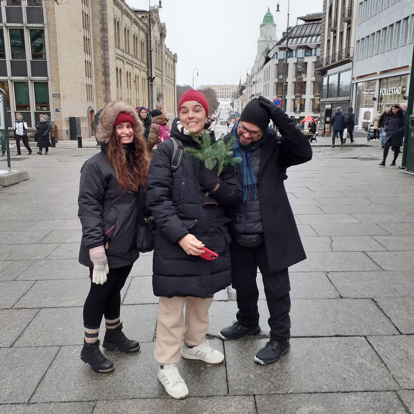 Three smiling OsloWay guests on Karl Johans gate in winter, with the Royal Palace in the background
