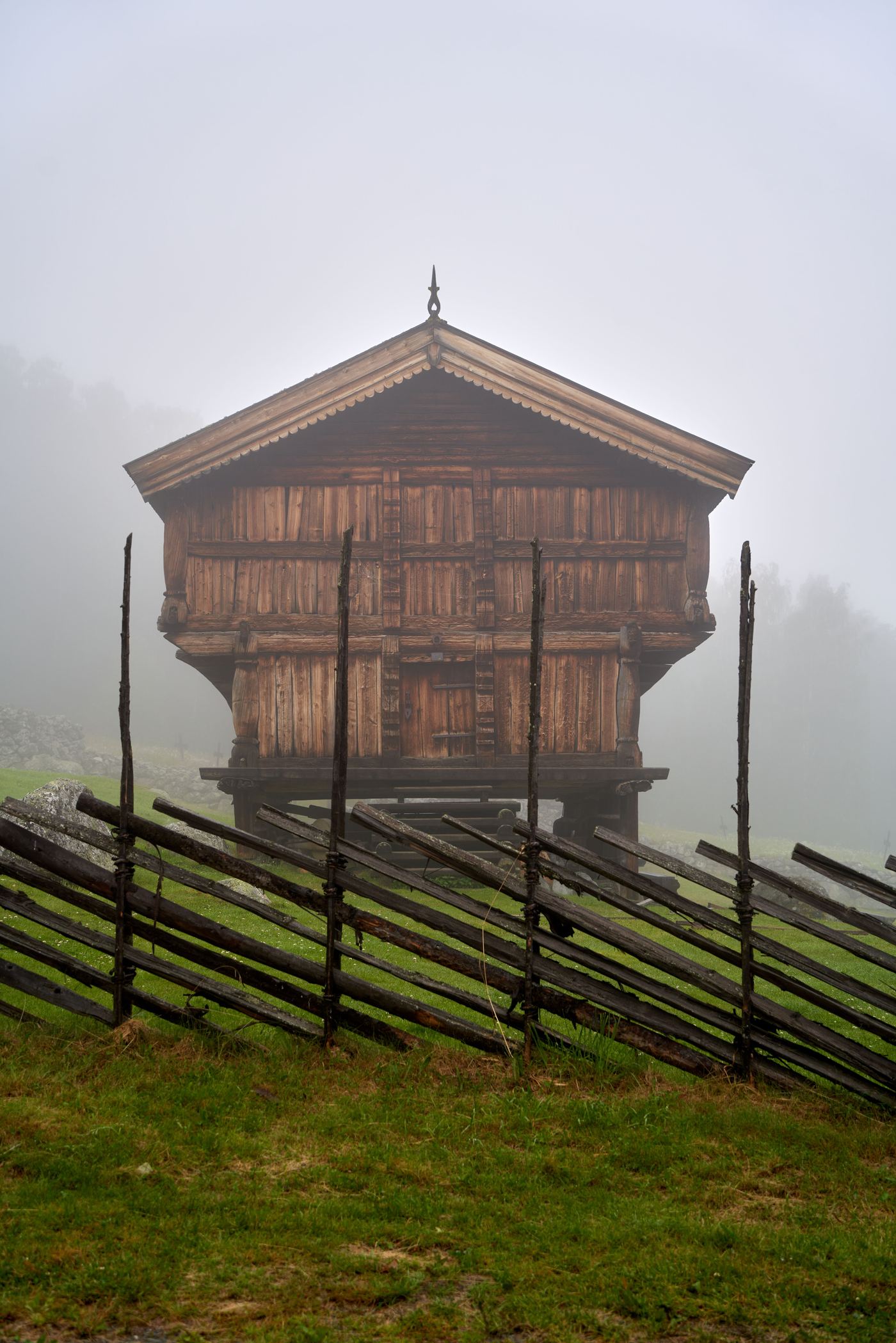 Traditional Norwegian stabbur storehouse on stilts standing in the mist behind an old wooden fence