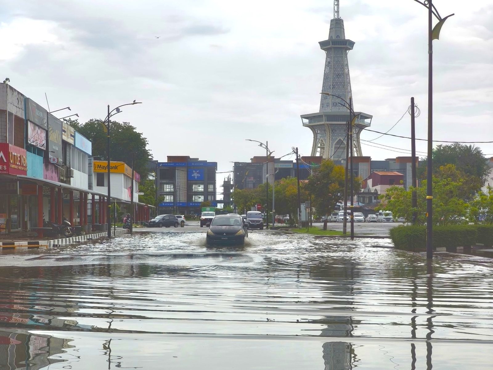 A car wading through floodwater covering a street in Kuah, with the MahaTower behind