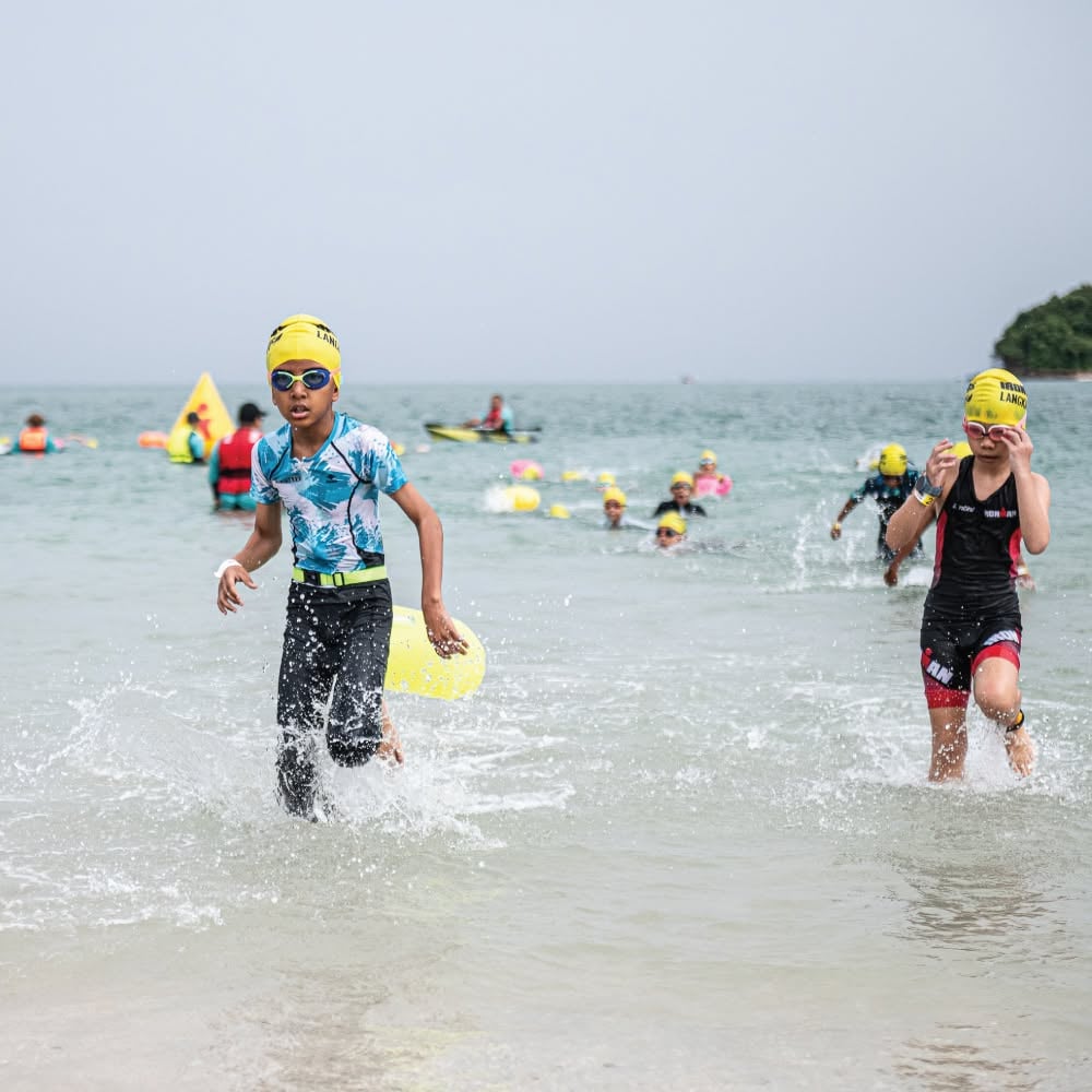 Young athletes running towards the IRONKIDS finish arch on the beach, with spectators behind the barrier