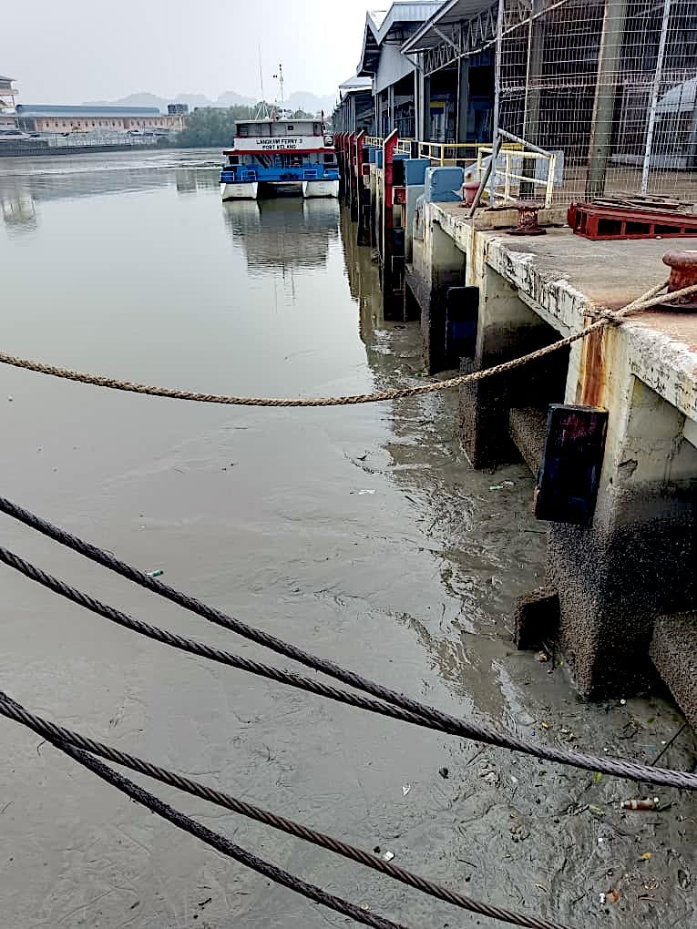 A ferry moored at a heavily silted Kuala Perlis jetty at low tide, with mud banks exposed along the berth