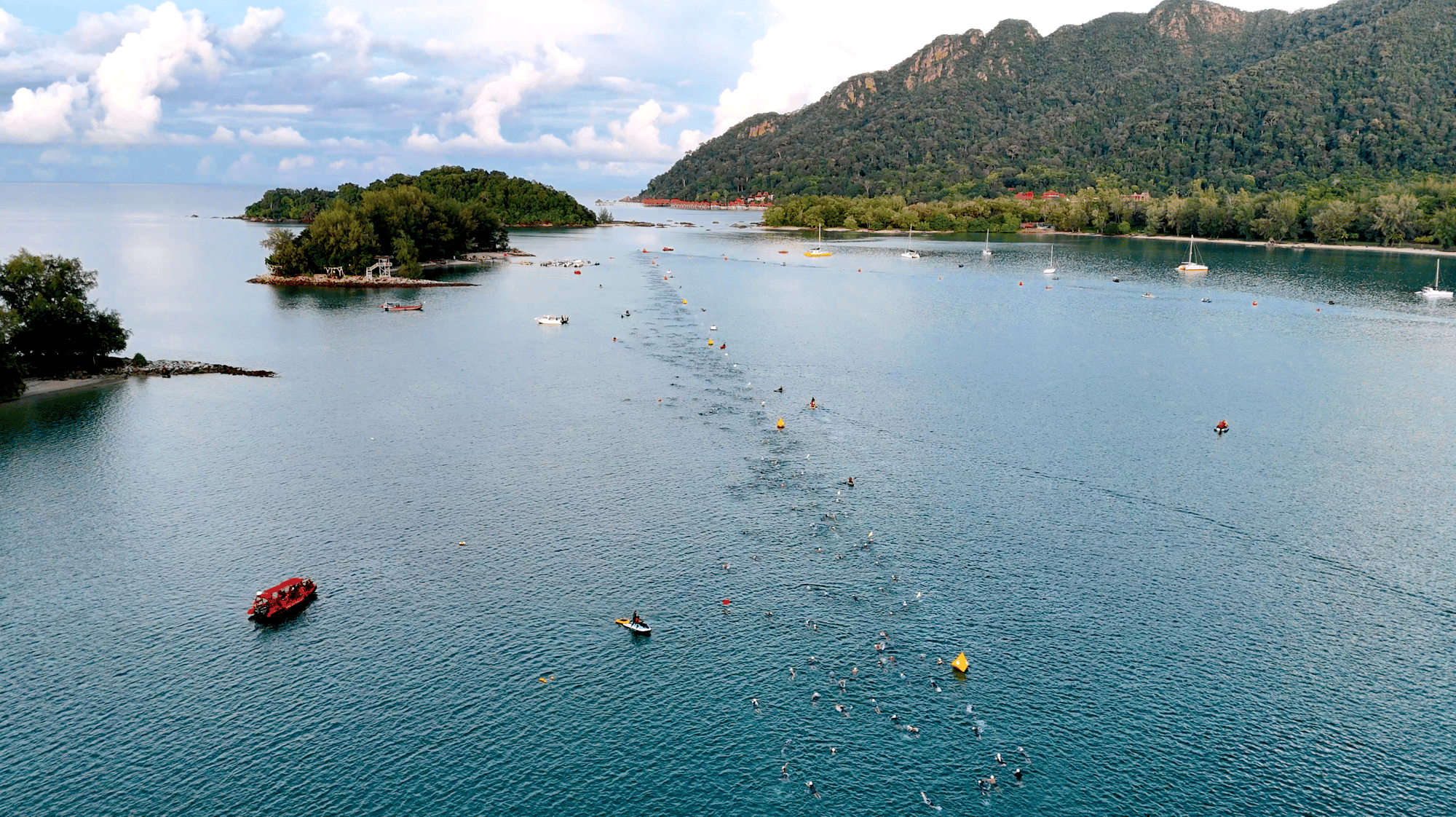 Aerial view of swimmers strung out along a line of buoys in sheltered water beside small islands, with rescue boats nearby