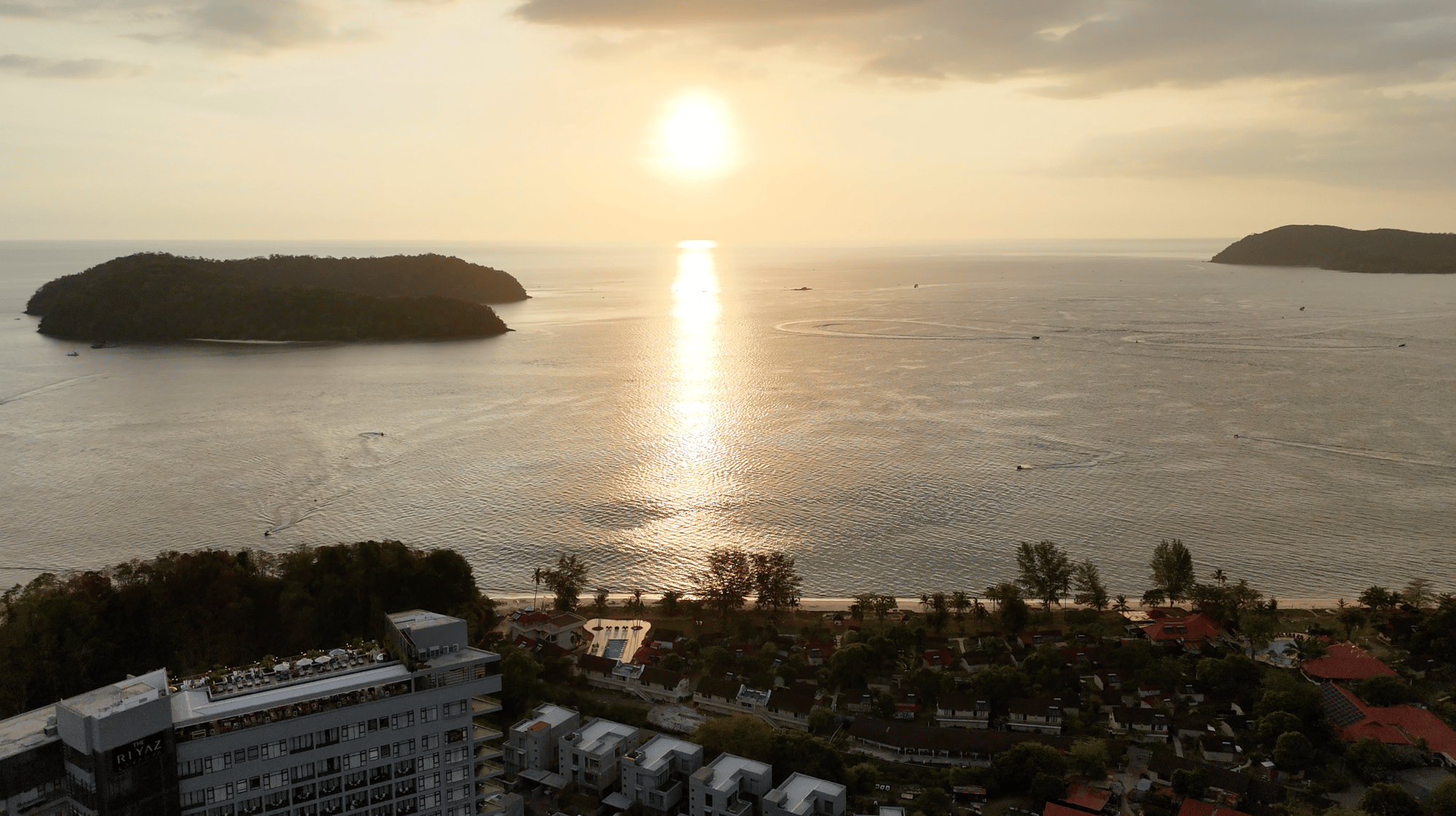 Aerial view of Pantai Tengah at sunset, the sun setting between offshore islands, with hotels and palms along the beach