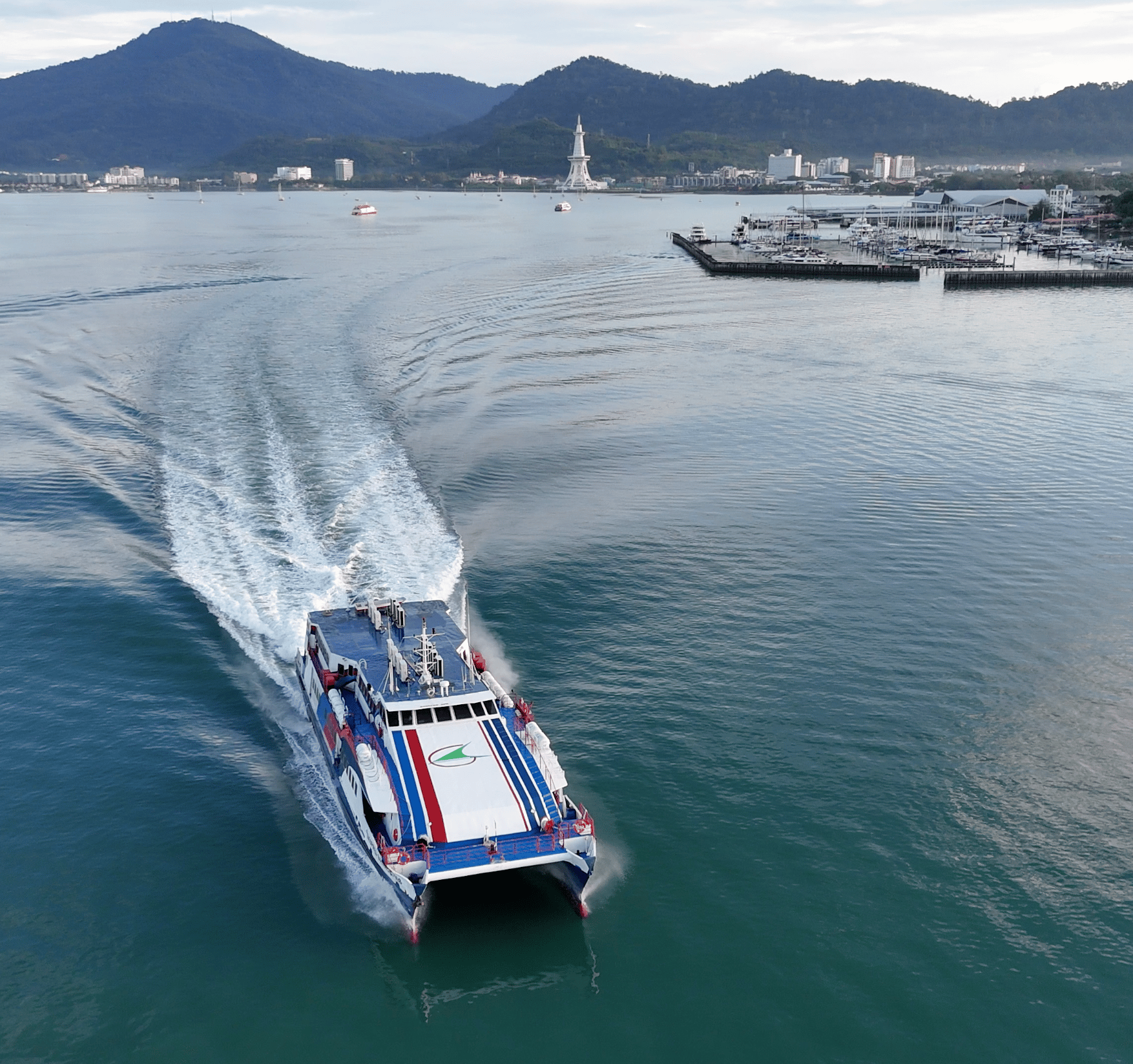 Aerial view of a passenger catamaran ferry approaching Kuah, with Eagle Square and the Langkawi skyline behind it