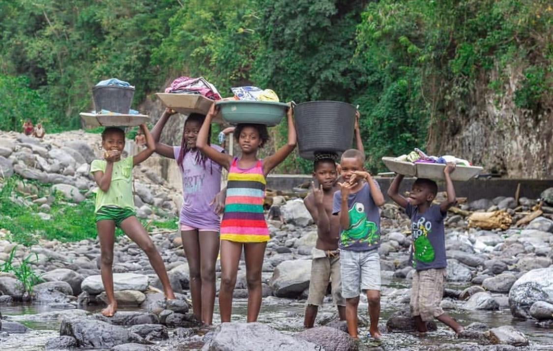 Crianças a transportar grandes bacias coloridas na cabeça ao longo de uma estrada em São Tomé