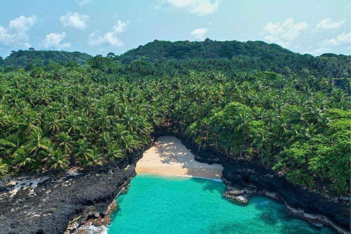 Aerial view of Praia Bateria on Ilhéu das Rolas, showing a secluded cove with turquoise water surrounded by black volcanic rocks and dense palm forest